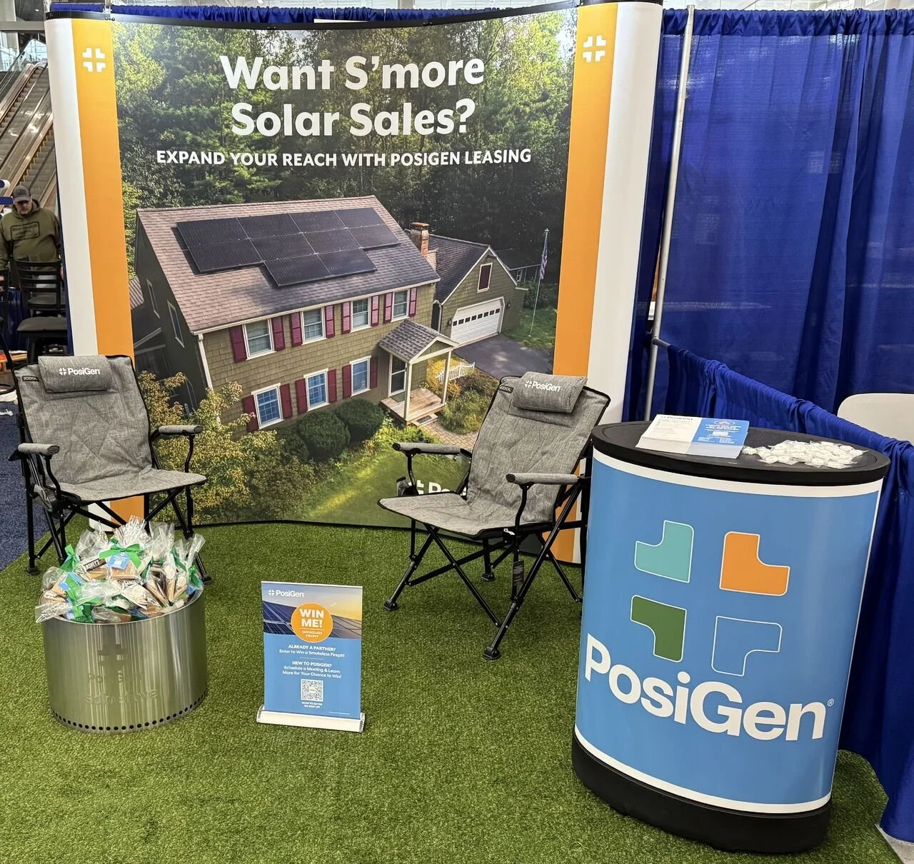 Booth for PosiGen company with a large backdrop featuring a house with solar panels on the roof. The booth has two gray outdoor chairs, a table with promotional items, and a PosiGen branded counter. The sign promotes solar sales and leasing options.