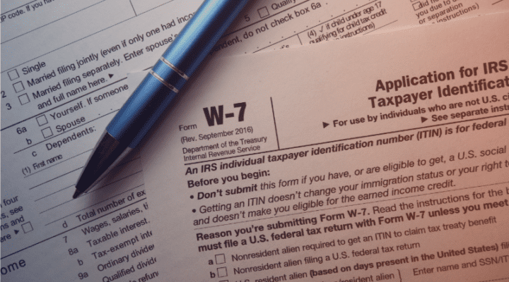 A close-up of a partially completed IRS Form W-7 with a blue pen resting on the paper and a checklist visible in the background.