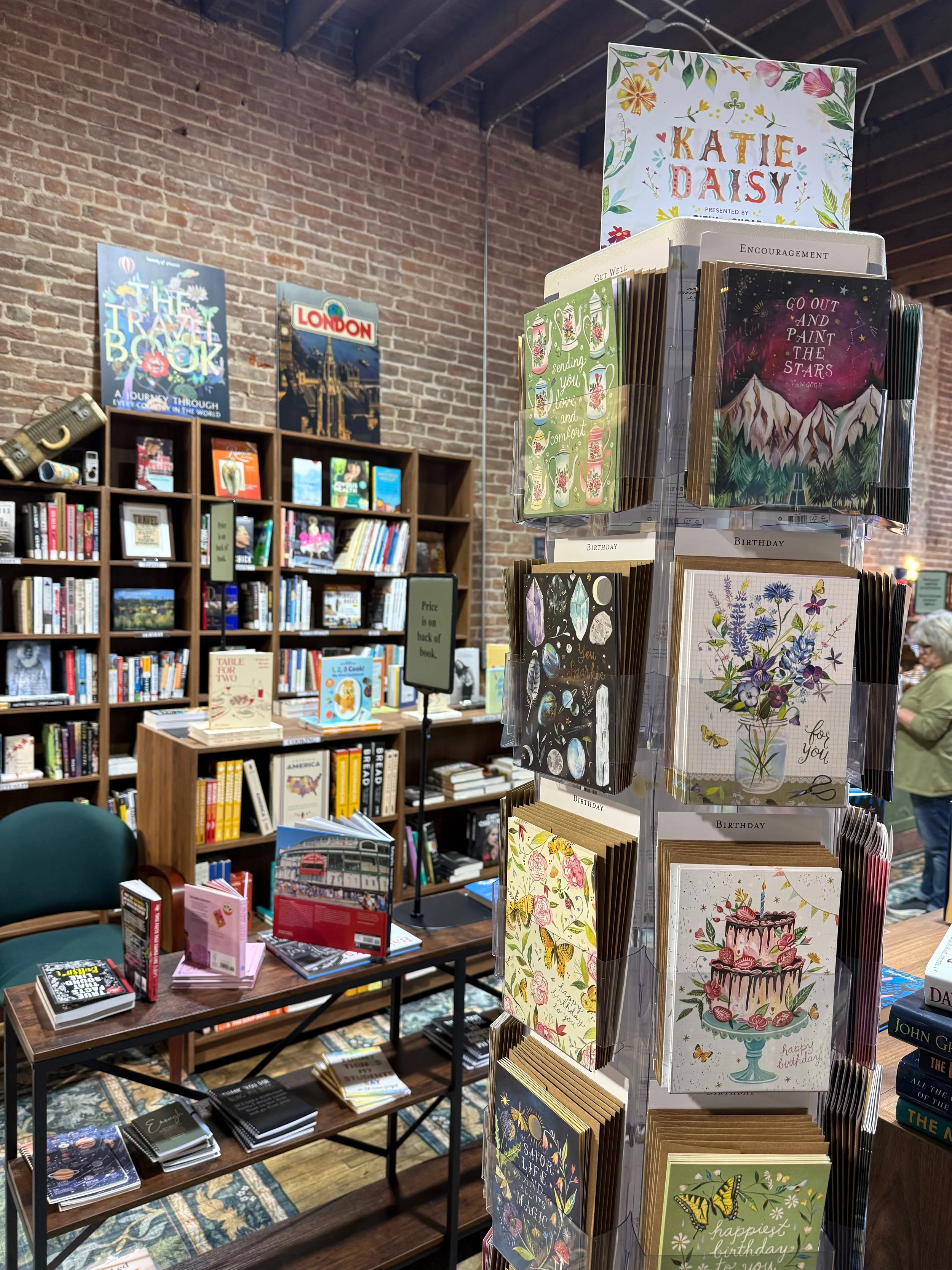 A display of greeting cards in a bookstore with a brick wall background and bookshelves filled with books.