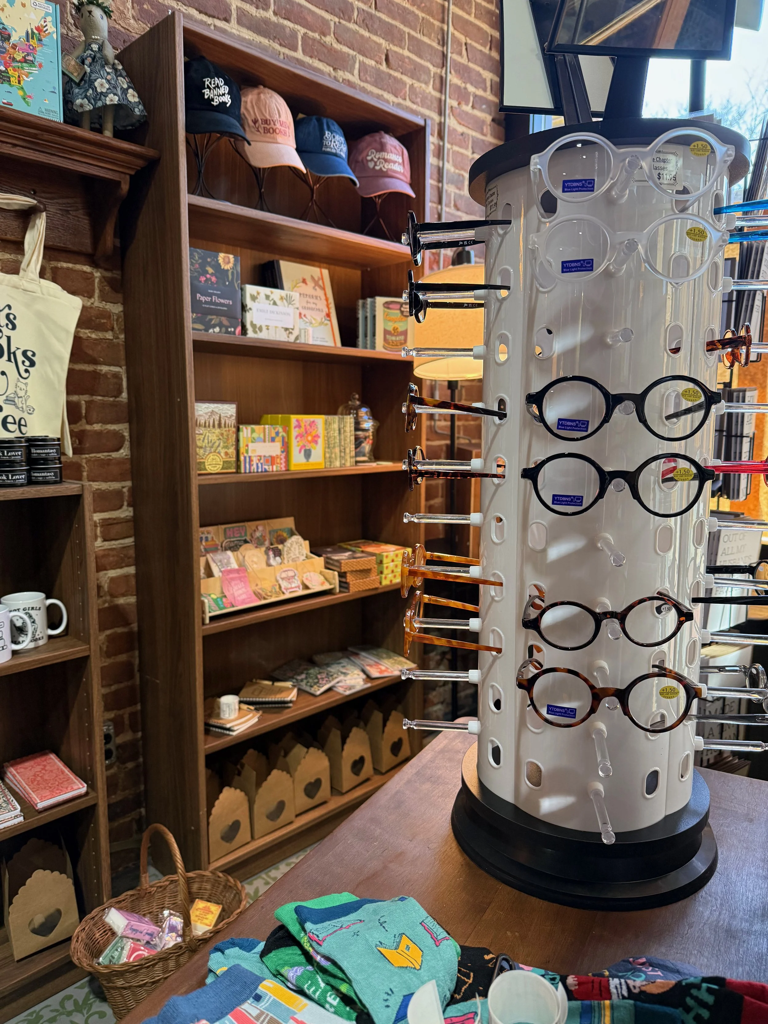 Display of eyeglasses on a rotating stand in a gift shop, with shelves of greeting cards, books, and hats in the background.