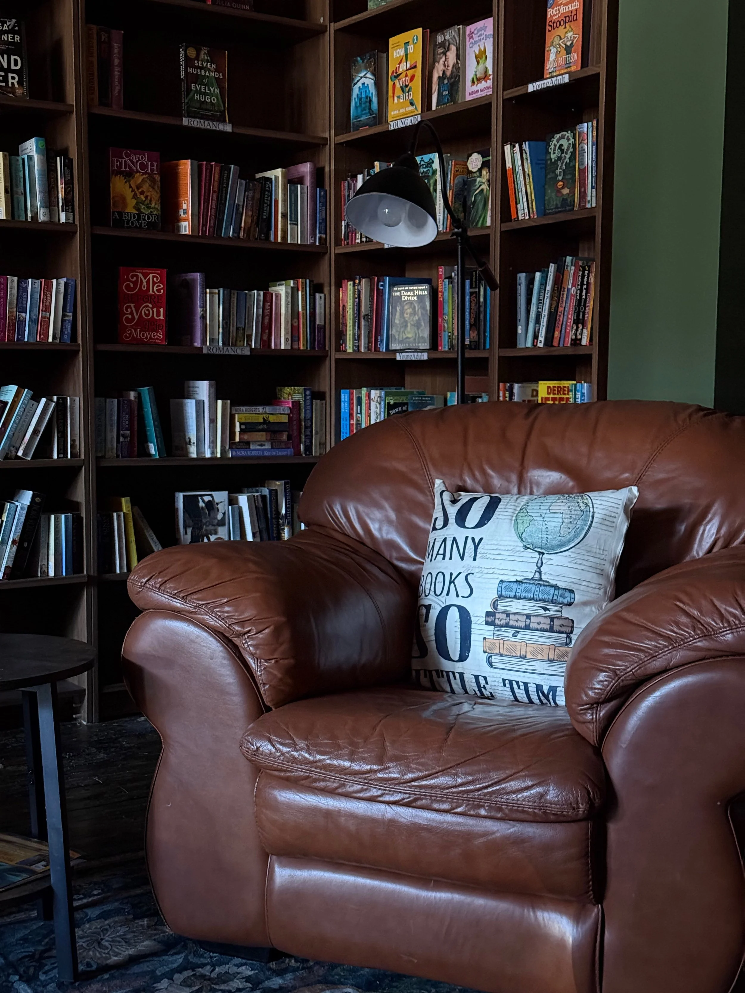 A cozy library corner with a leather armchair, a decorative pillow that reads 'So many books, so little time,' a bookcase filled with books, and a reading lamp.