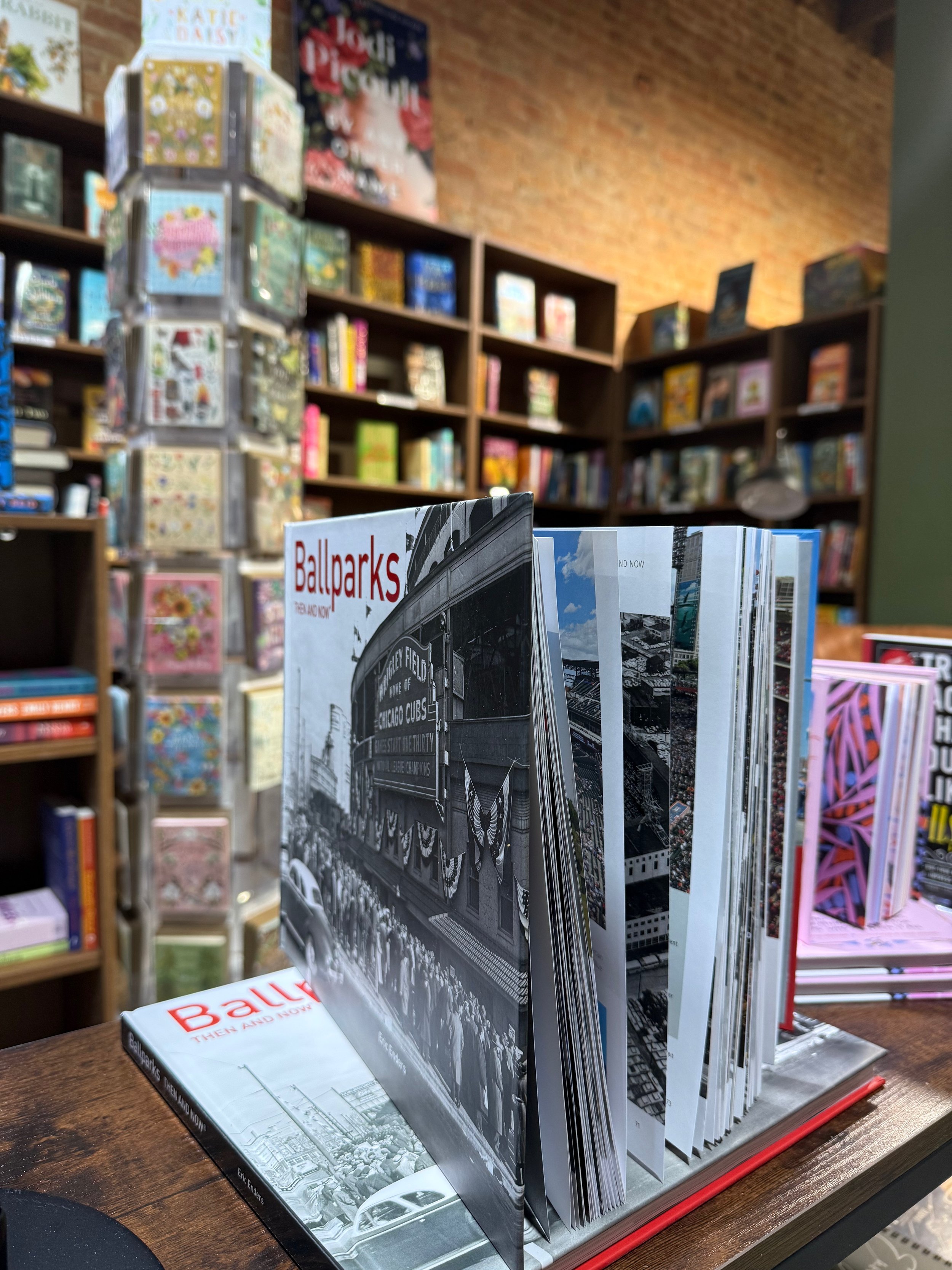 A display of Chicago-related books, including a black-and-white book titled 'Ballparks' and an open book with black-and-white images, on a wooden table in a bookstore with shelves of colorful books in the background.