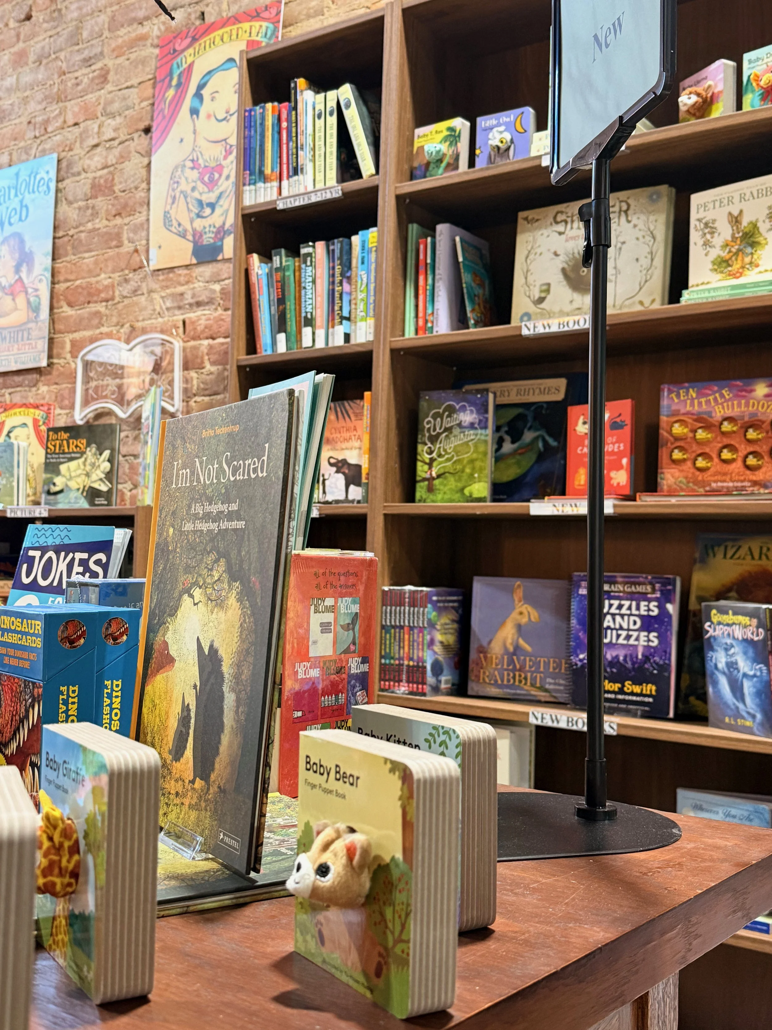 A cozy bookstore with wooden shelves filled with children's books. In the foreground are small plush animal bookmarks labeled 'Baby Giraffe' and 'Baby Bear.' The background shows colorful book covers and posters on the brick wall.