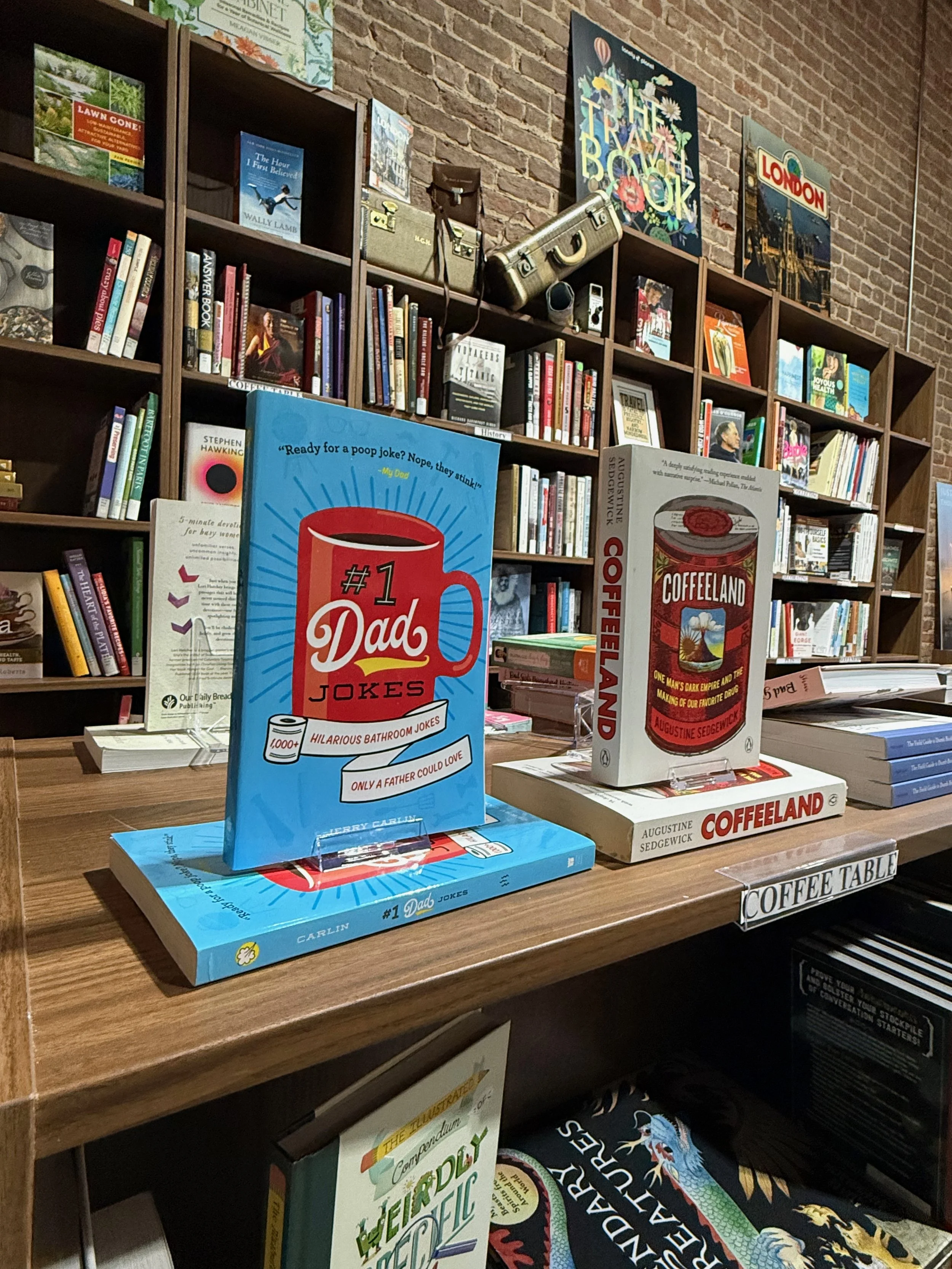 Books on a wooden table, including one titled '#1 Dad Jokes' and another titled 'Coffeeland,' with a shelf of books and decorative posters on a brick wall in the background.