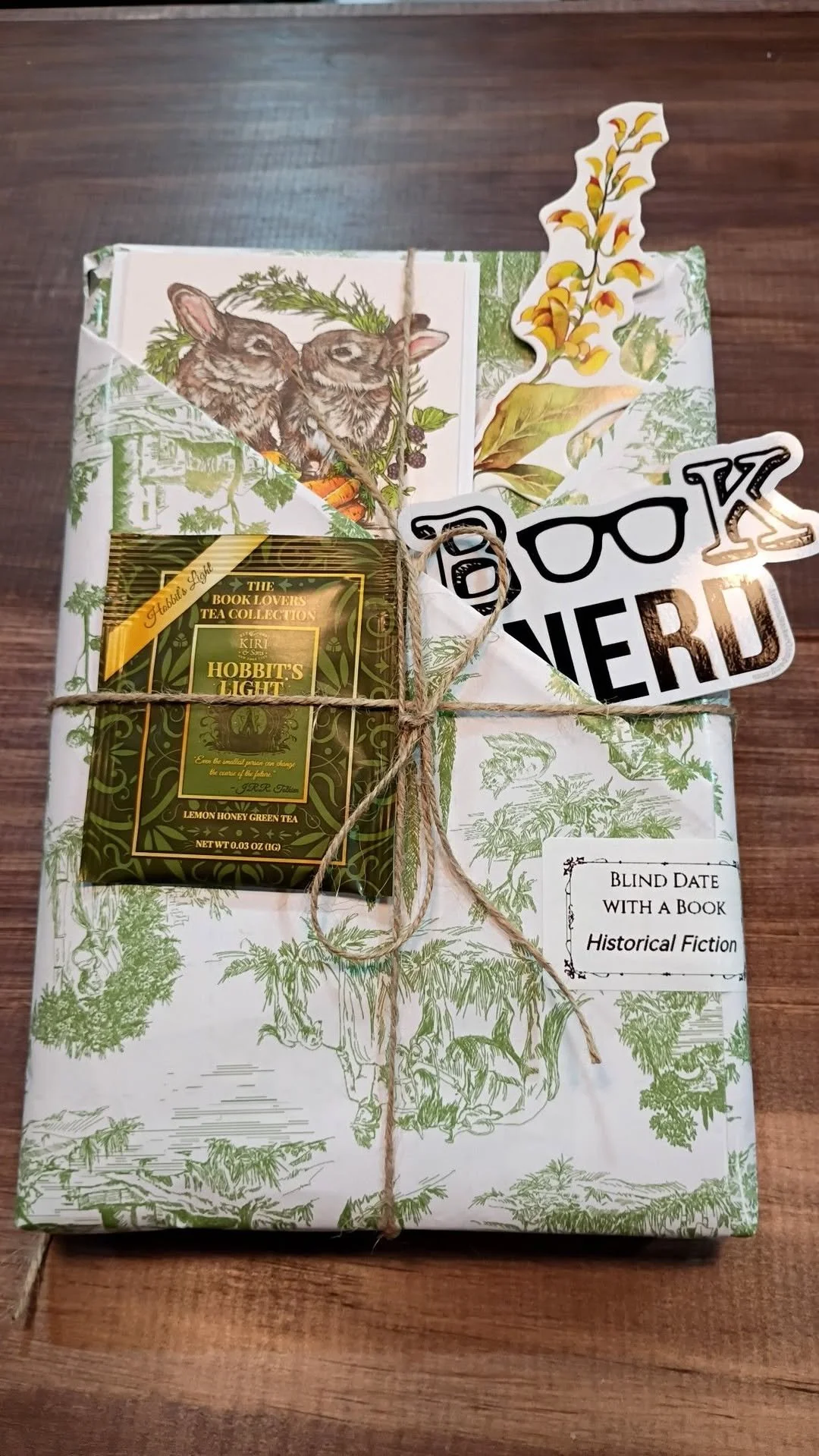 A wrapped gift with green and white book-themed paper, tied with twine, includes a box of Hobbit's Light lemon honey green tea, with notes and stickers attached.