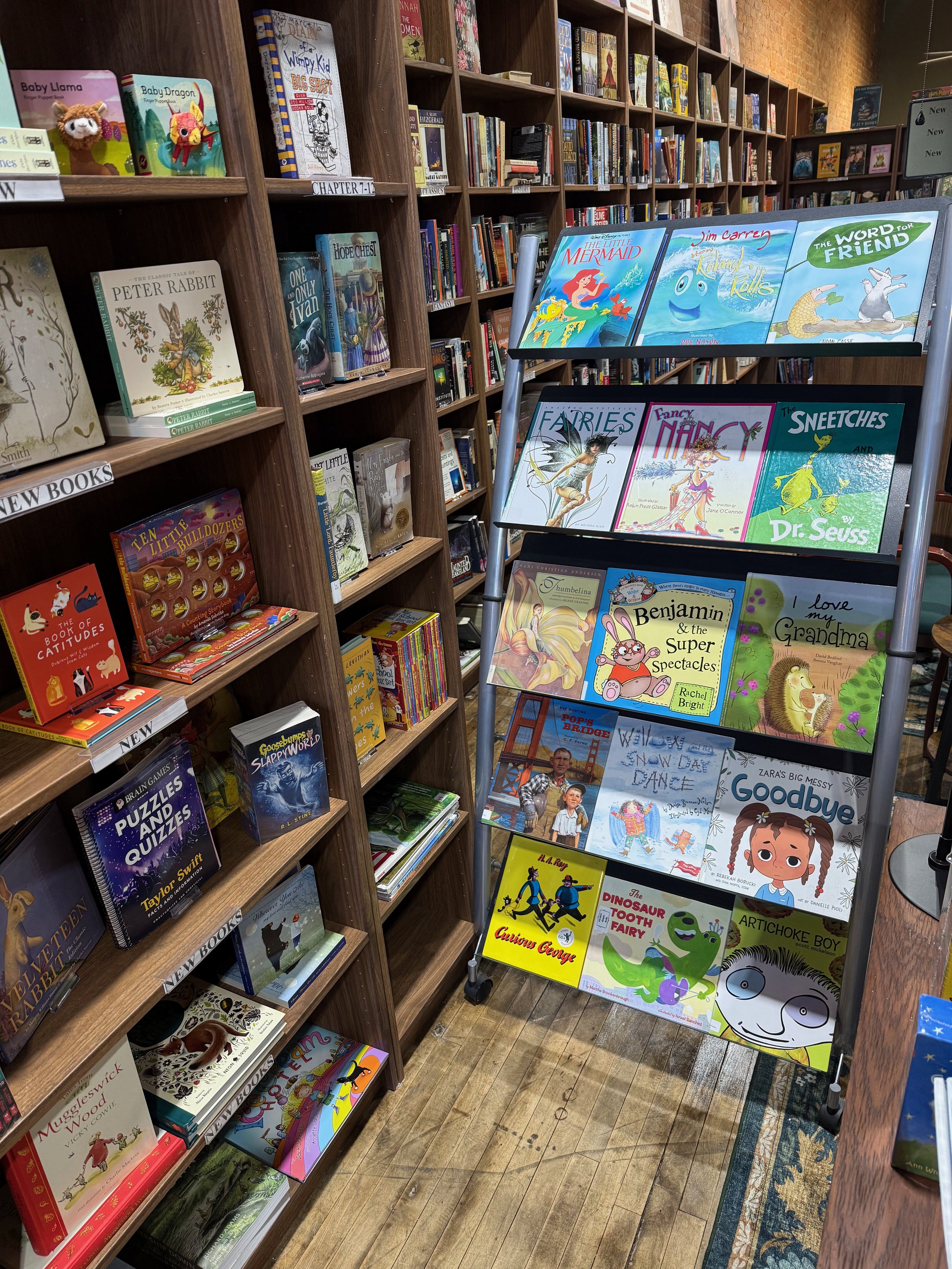 A bookstore display with children's books on a tiered stand, featuring titles like 'The Little Mermaid,' 'Fancy Nancy,' and 'Goodbye,' surrounded by bookshelves filled with various books.