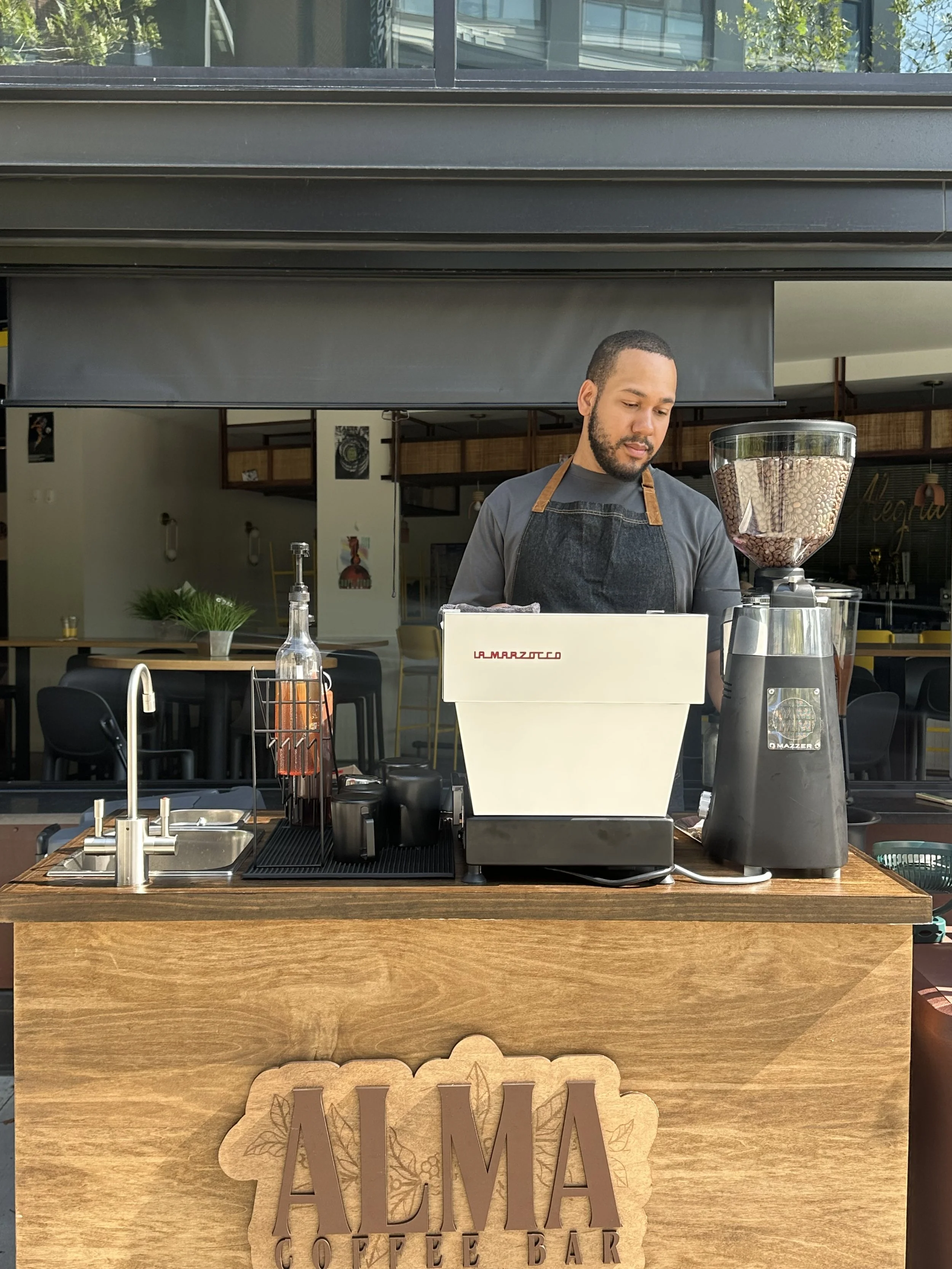 A barista preparing coffee at Alma Coffee Bar, with coffee beans in a grinder and a small sink on a wooden counter, inside a modern cafe.