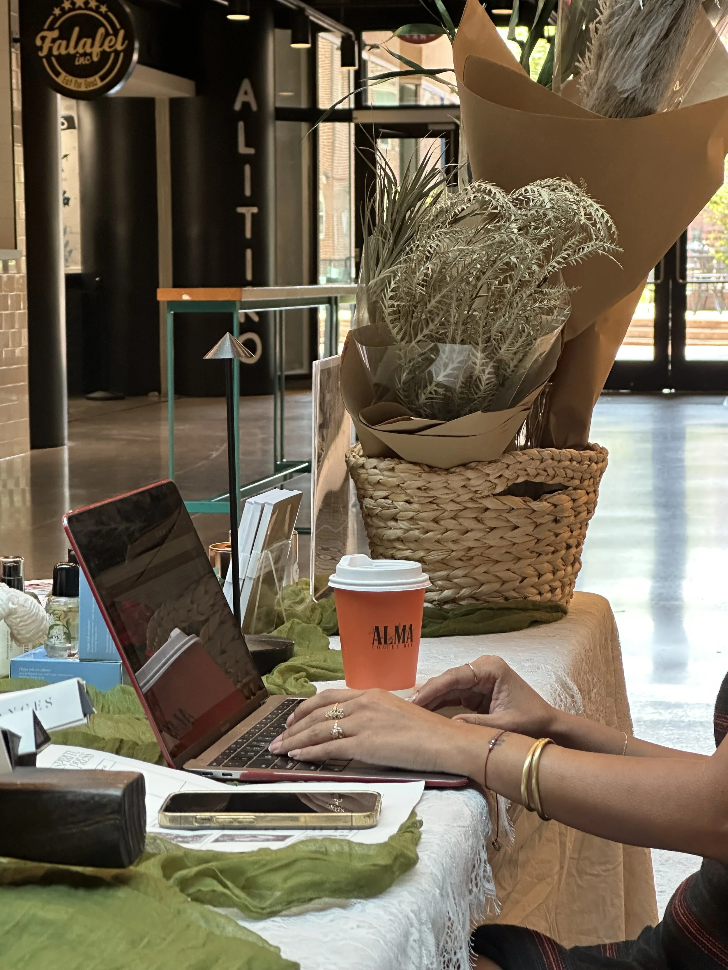 A person working on a laptop at a table with a large potted plant wrapped in paper, a disposable coffee cup, and other items including a phone, notebooks, and decorative objects. There are signs and windows in the background.
