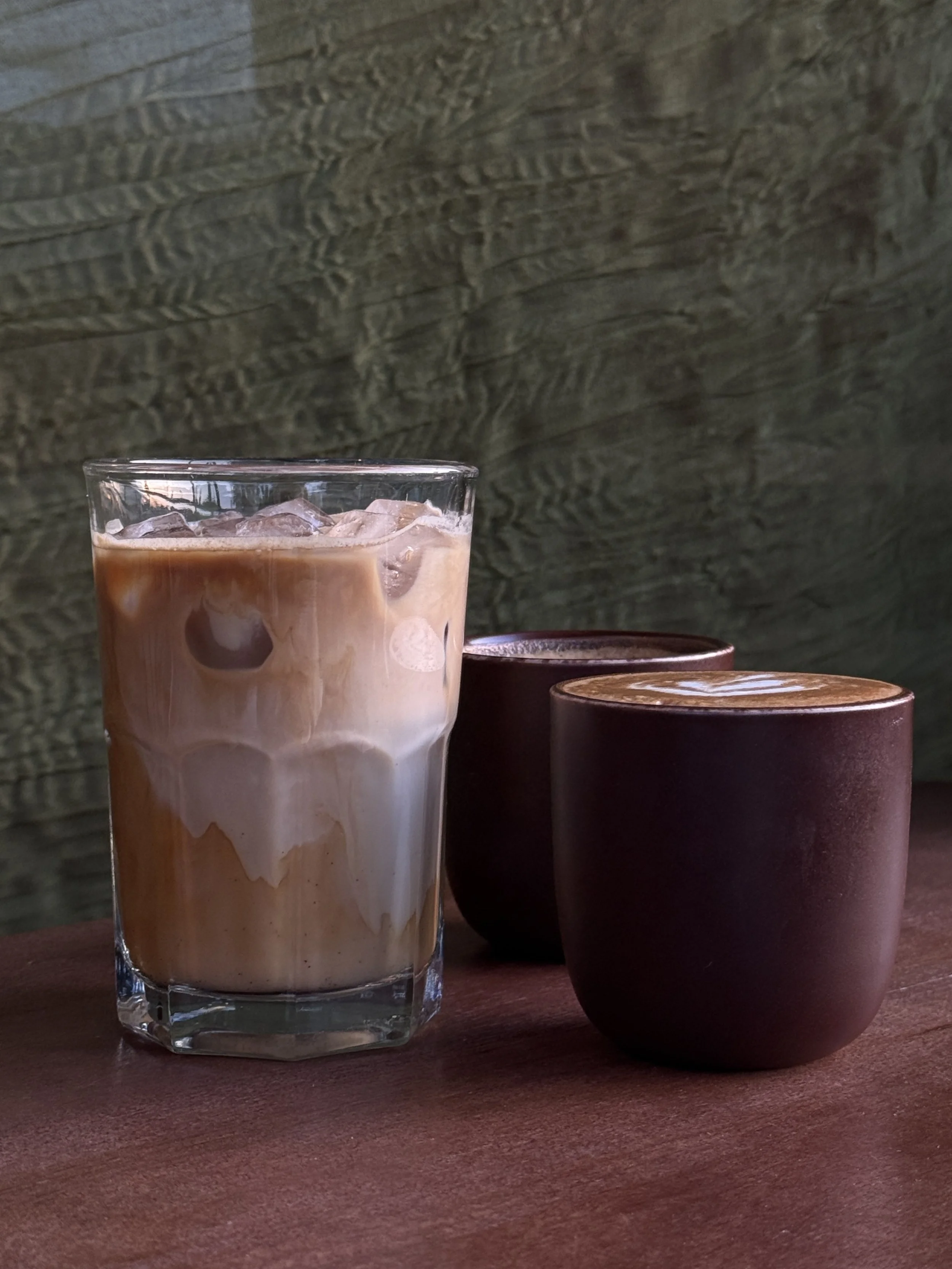 A tall glass of iced coffee with milk on a wooden table, with two small dark ceramic cups beside it, against a rustic wooden wall background.