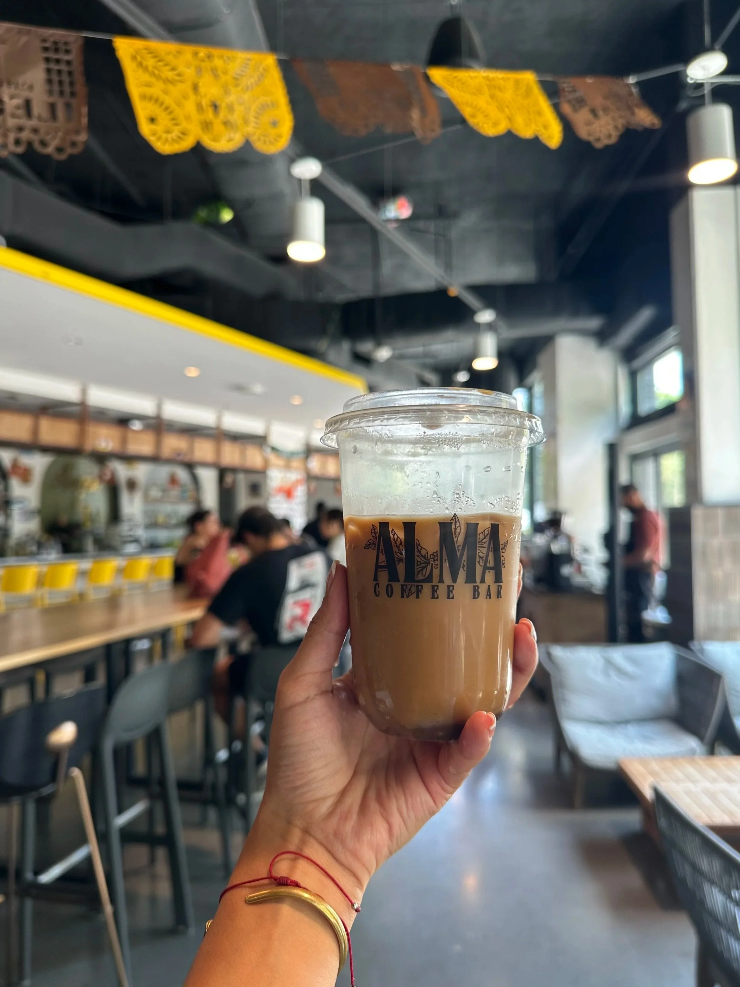 Hand holding a plastic cup of iced coffee with the logo 'ALMA Coffee Bar' in a coffee shop with seating and hanging decorations