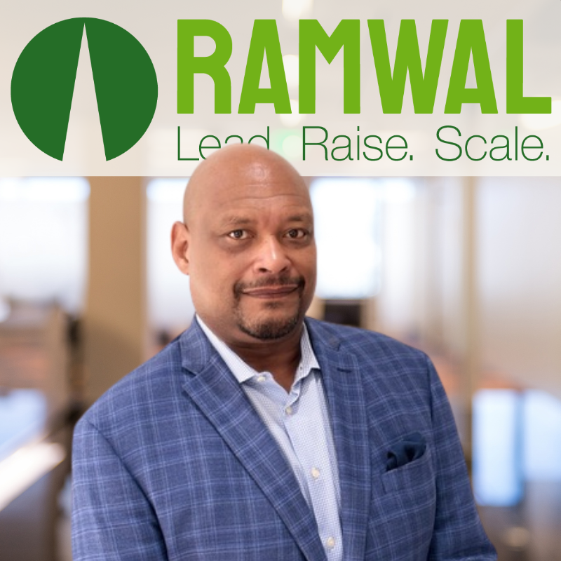A man in a blue checkered blazer and light blue shirt standing indoors, with a banner above him displaying the Ramwal logo and the words 'Lead. Raise. Scale.'