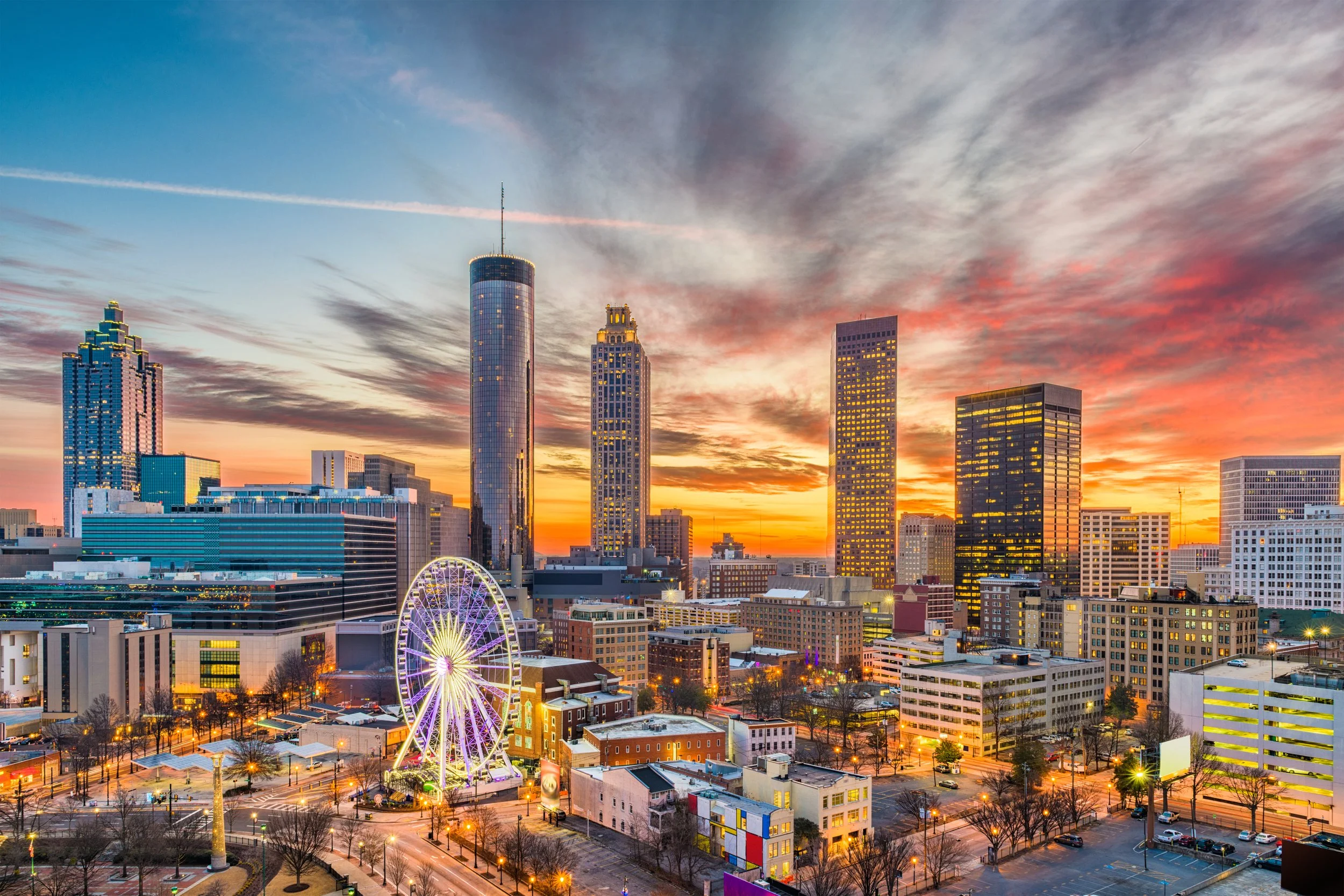 A city skyline at sunset featuring tall skyscrapers, a Ferris wheel, and illuminated streets in Atlanta.