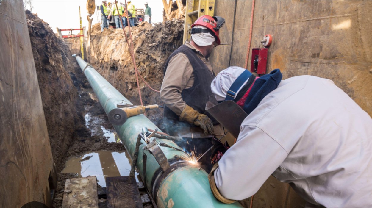 Construction workers repairing a large pipe in a trench, with one worker welding and others supervising.