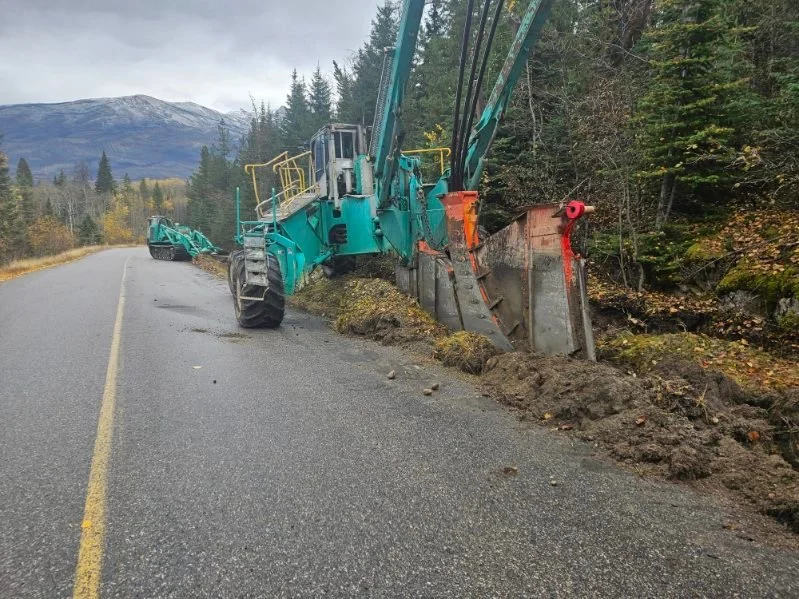 A large blue excavator is working on the side of a mountain road, with part of the road washed out and soil displaced along the edge. The scene is set in a forested area with mountains visible in the background.