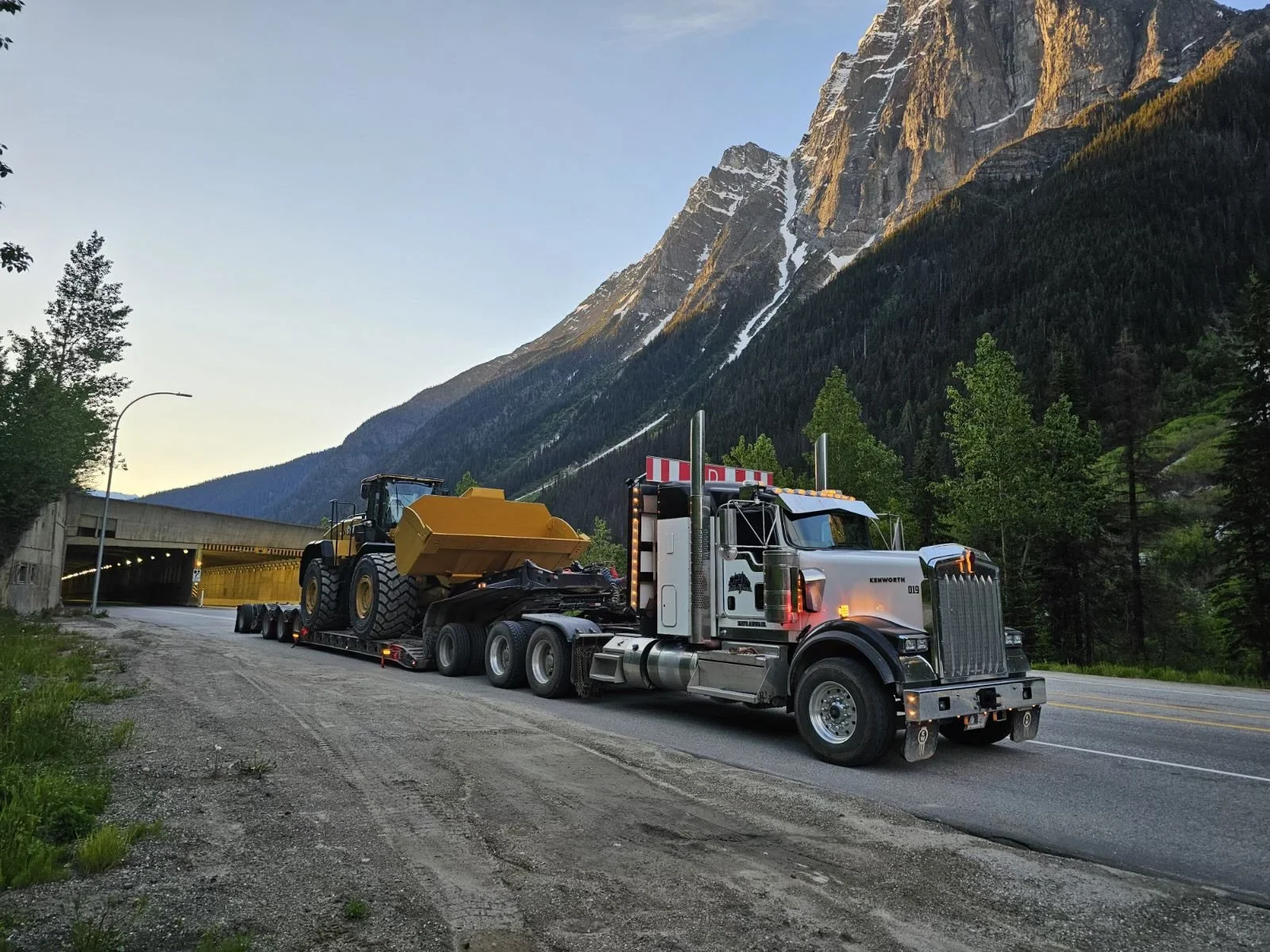 A semi-truck with a large yellow construction vehicle on a flatbed, parked on the side of a mountain road with forested mountains and a tunnel in the background.