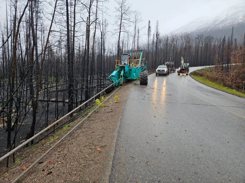 A tree trimming machine working on a roadside that has been affected by a wildfire, with charred trees on the left and a wet road curving to the right in the background.