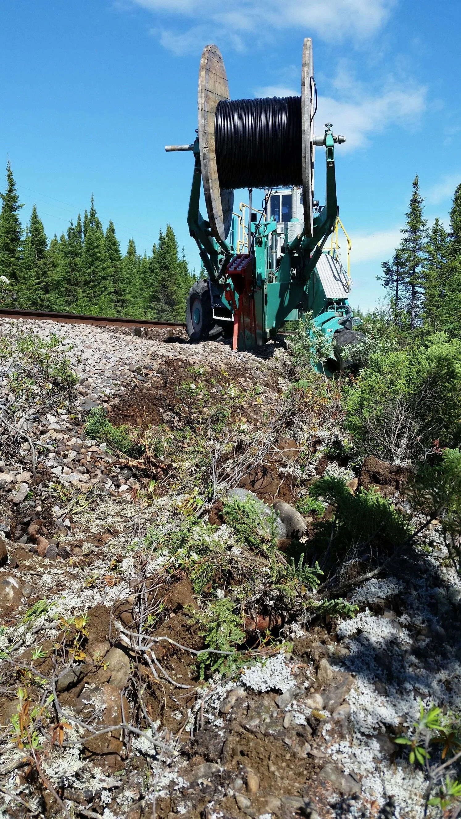 A large green forest maintenance machine with a spool of black cable, situated next to a railway track, outdoors amidst trees and vegetation.