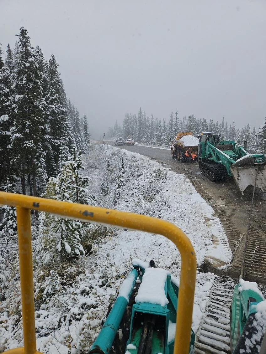 A snowy mountain landscape with trees covered in snow and a road being cleared by construction machinery, including a green snowplow and an orange snow removal vehicle, with four-wheel drive vehicles on the road.