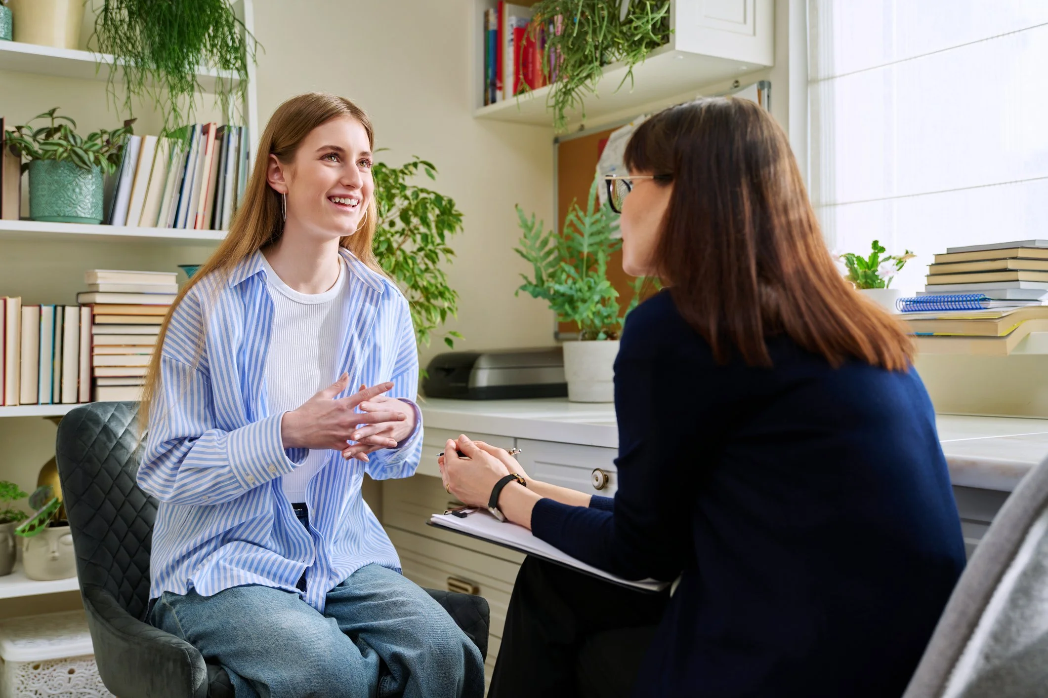 A young woman in a light blue striped shirt sitting and smiling during a conversation with a counselor or therapist, who has brown hair and glasses, in an office decorated with books and plants.