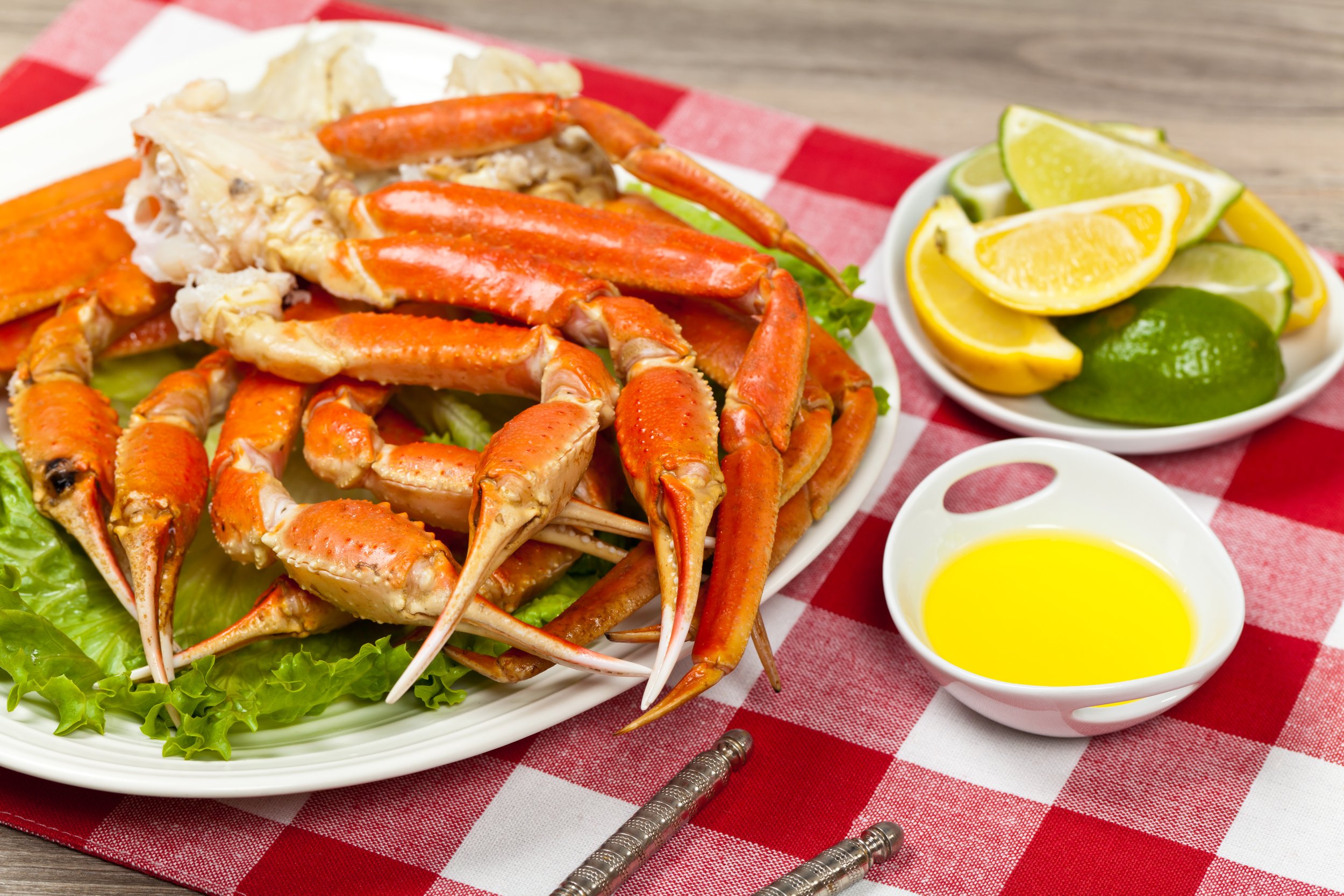 Crab legs served on a bed of lettuce with lemon and lime wedges, lemon butter sauce, on a red and white checkered tablecloth.