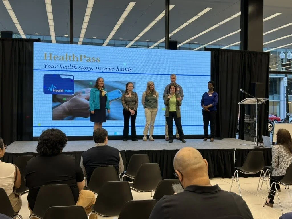 Christina Dzingala, Diana Finegold, Emily Modde, Zeina Shreif, Greg Winston, and Yvonne Paintsil standing on stage addressing questions from the audience about their project HealthPass. standing on stand presenting their project.