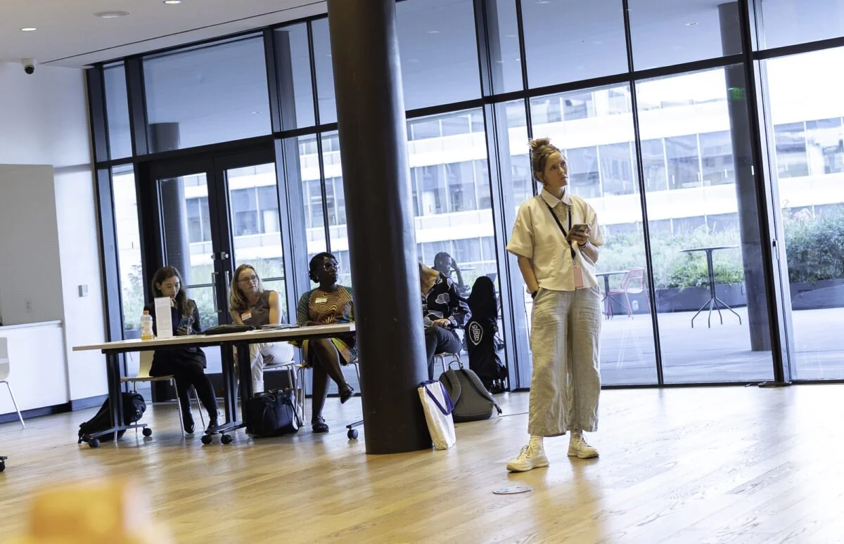 Chelsea Kirkland standing in a room at the DC Public Library with four women sitting at a table behind her.