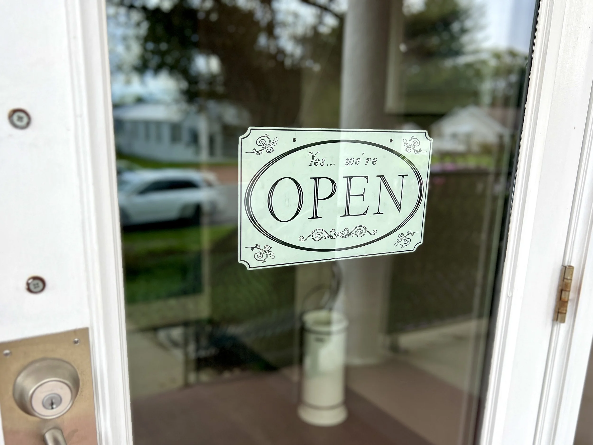Glass door with a white sign that says, 'Yes... we're OPEN' in black lettering. Reflection of trees and houses outside. Part of a door lock and handle visible on the left side.