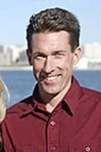 Portrait of a man with dark hair, smiling, wearing a maroon shirt, outdoors near water with city buildings in the background.