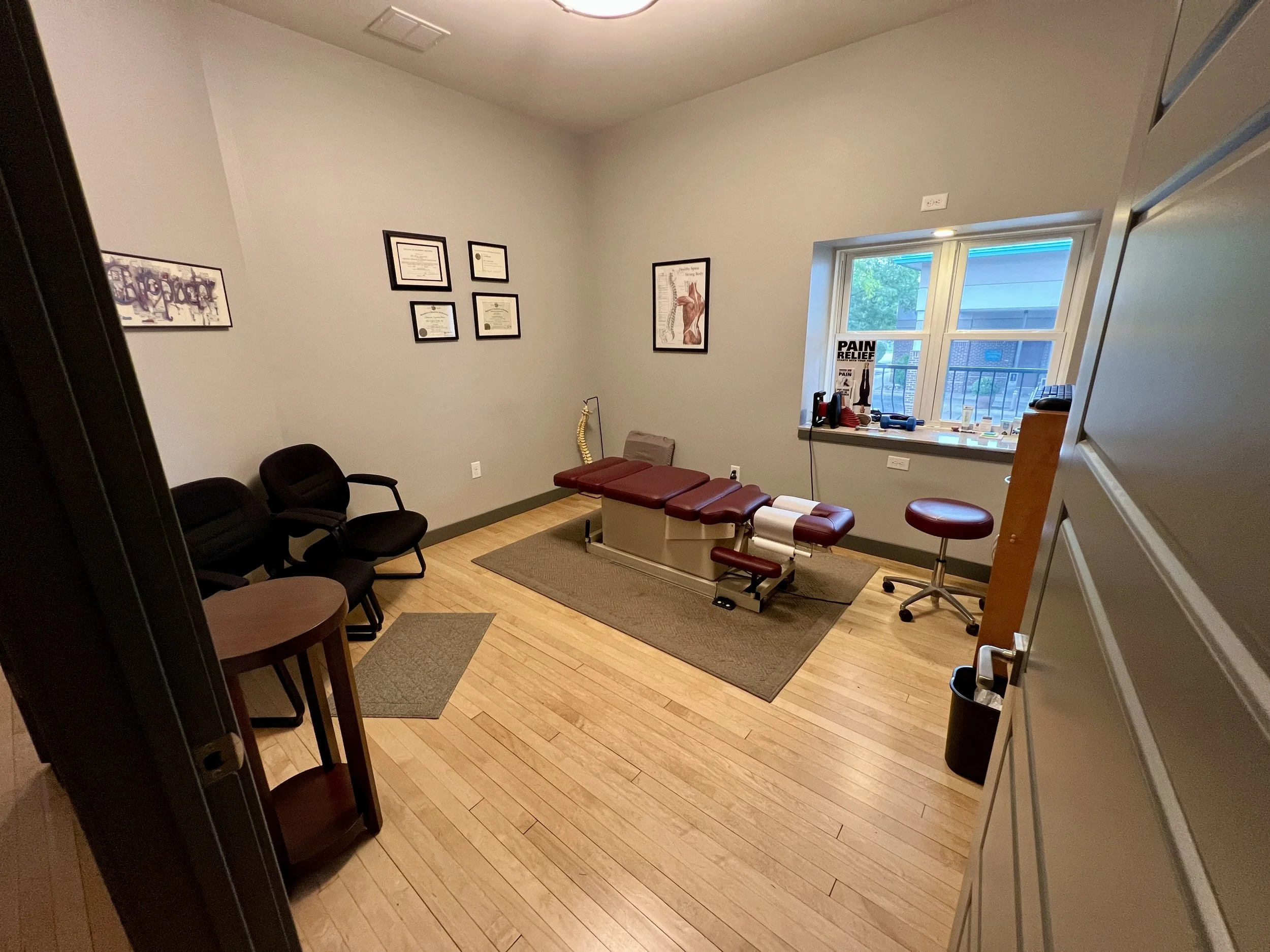 A chiropractic or medical examination room with examination table, framed certificates and posters on the walls, chairs, a window with a view of neighboring buildings, and various medical supplies on the windowsill.