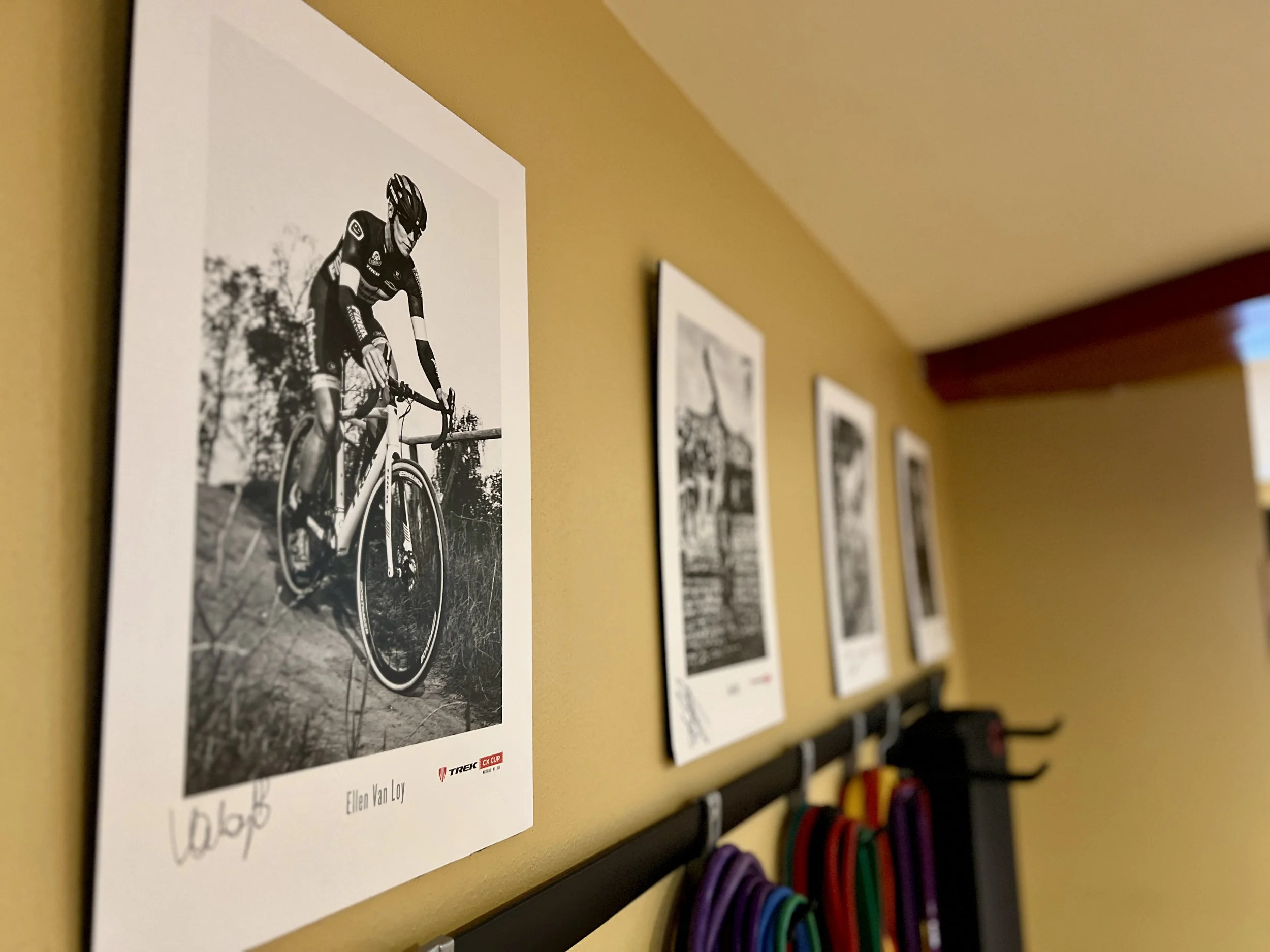 Verona Fitness Studio artwork black and white photograph of a cyclist riding a bike, displayed on a yellow wall among other framed photographs in a gallery or exhibit setting.
