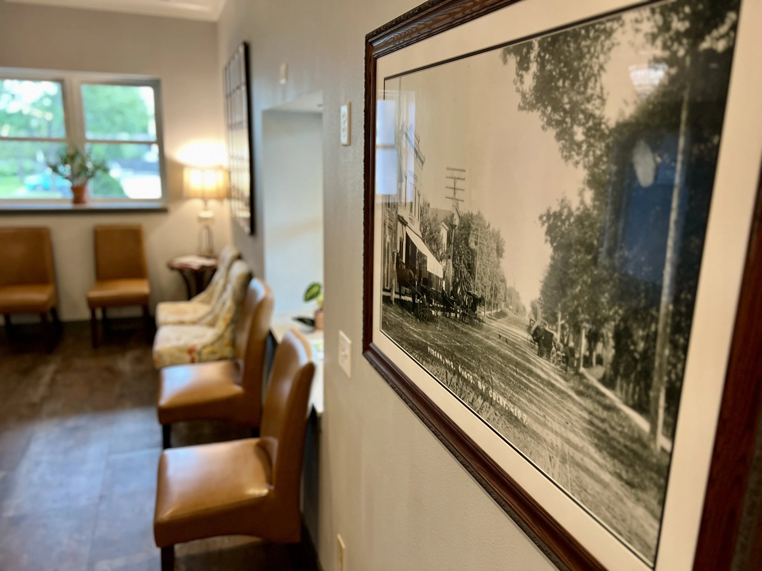 Interior of a room with a framed photograph of a street scene on the wall, wooden chairs, a window with a potted plant, a lamp, and upholstered chairs.