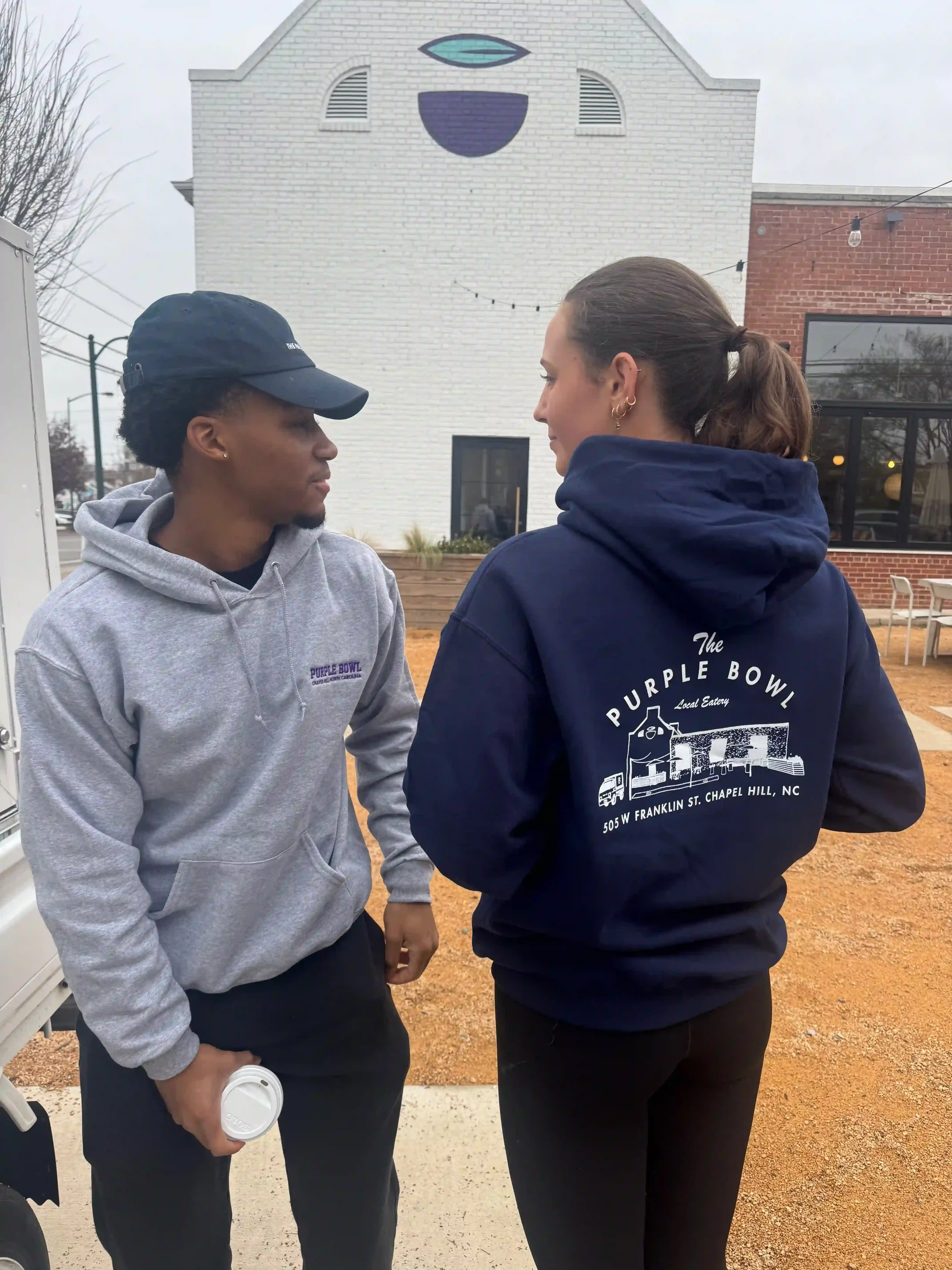 A man and woman look at each other wearing Purple Bowl hoodies. The man is wearing a heather gray hoodie with The Purple Bowl printed on it, and the woman is wearing a navy hoodie with The Purple Bowl's building graphic on the back.