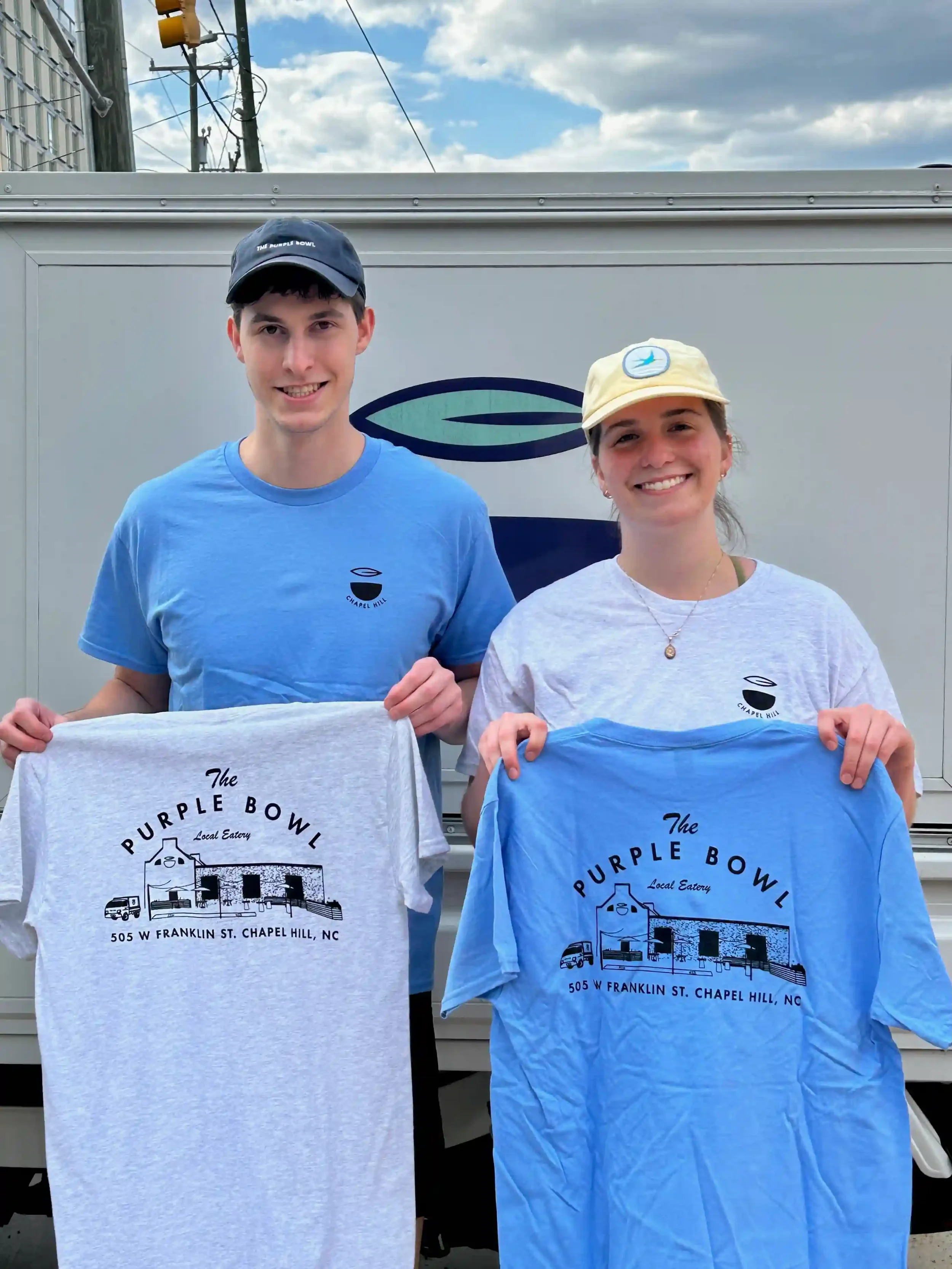 Two smiling people holding T-shirts with the logo and address of The Purple Bowl, a local eatery in Chapel Hill, NC, standing in front of a light-colored vehicle.