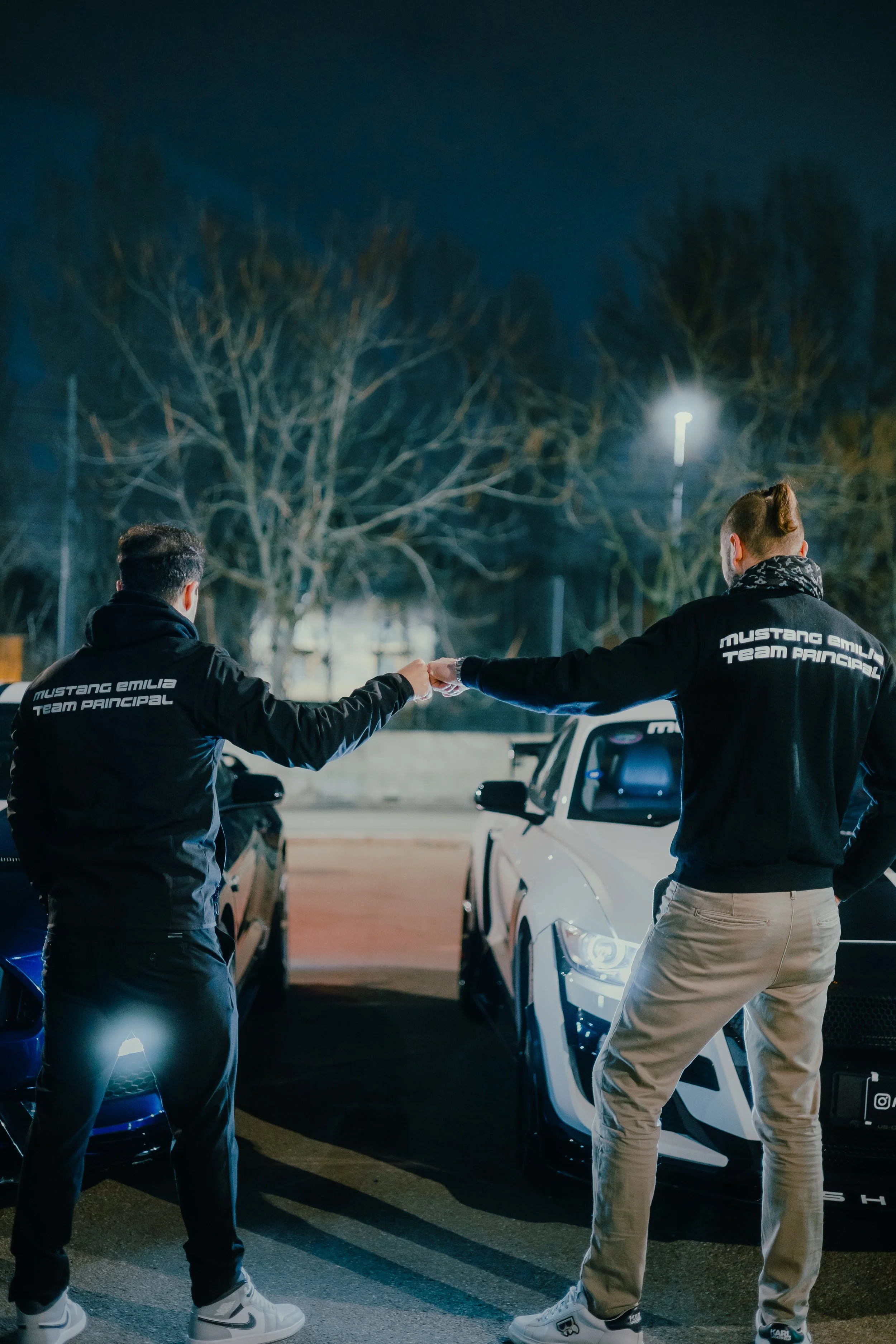 Due uomini con giubbotti con scritto 'Mustang Emilia Team Principal' si scambiano un oggetto all'aperto di sera, tra auto sportive.