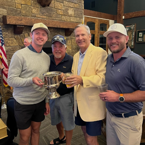 Four men standing together at an indoor event, smiling, with a large trophy and a drink in hand.