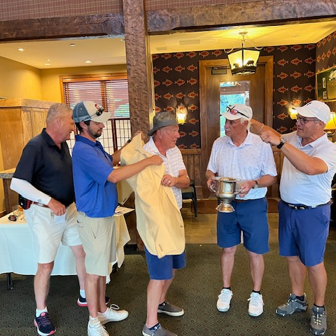 Six men in golf attire standing inside a restaurant or clubhouse, engaging in conversation and celebrating, with one holding a trophy.