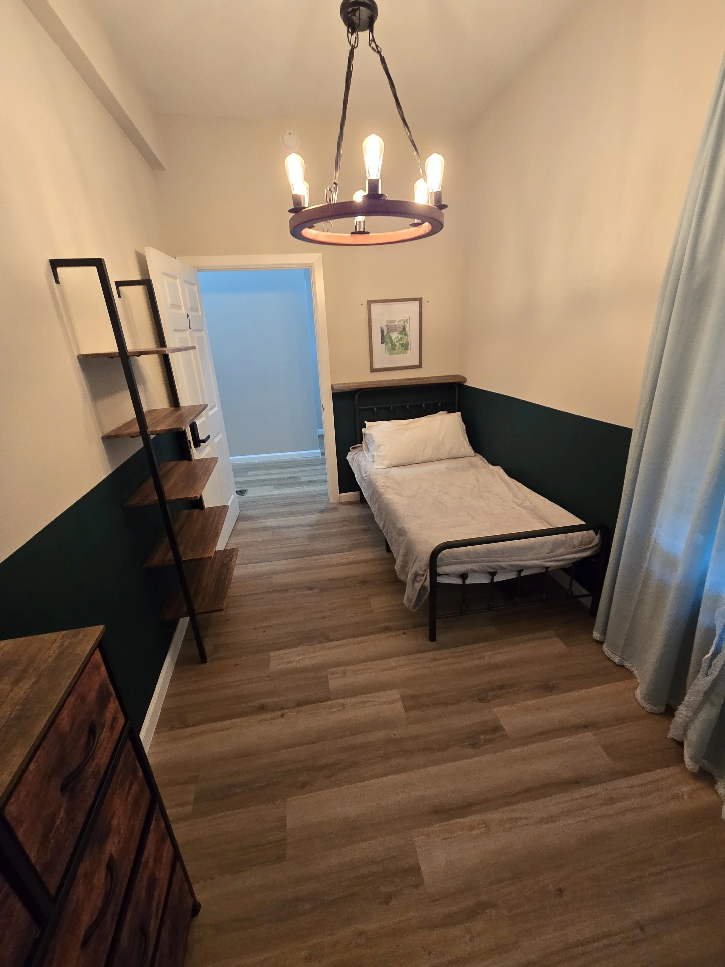 A small bedroom with a single bed and a dark metal headboard, a wooden shelf on black metal frame, a dresser, and a framed picture on the wall, illuminated by a chandelier with amber bulbs. The floor is wood, and there's a window with light curtains 