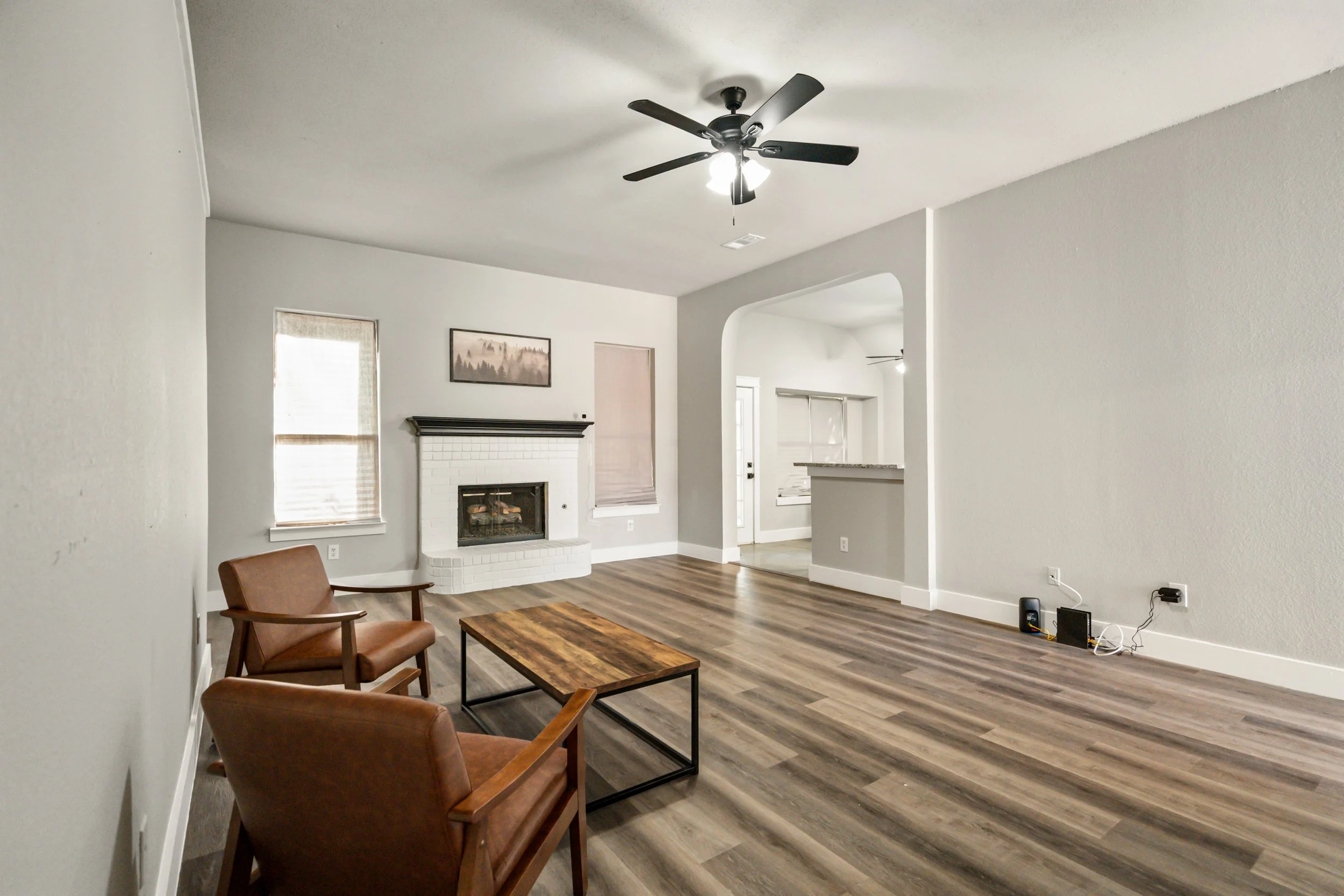 Empty living room with wood flooring, white walls, a ceiling fan, a fireplace, a window with blinds, and two brown chairs near a wooden coffee table.