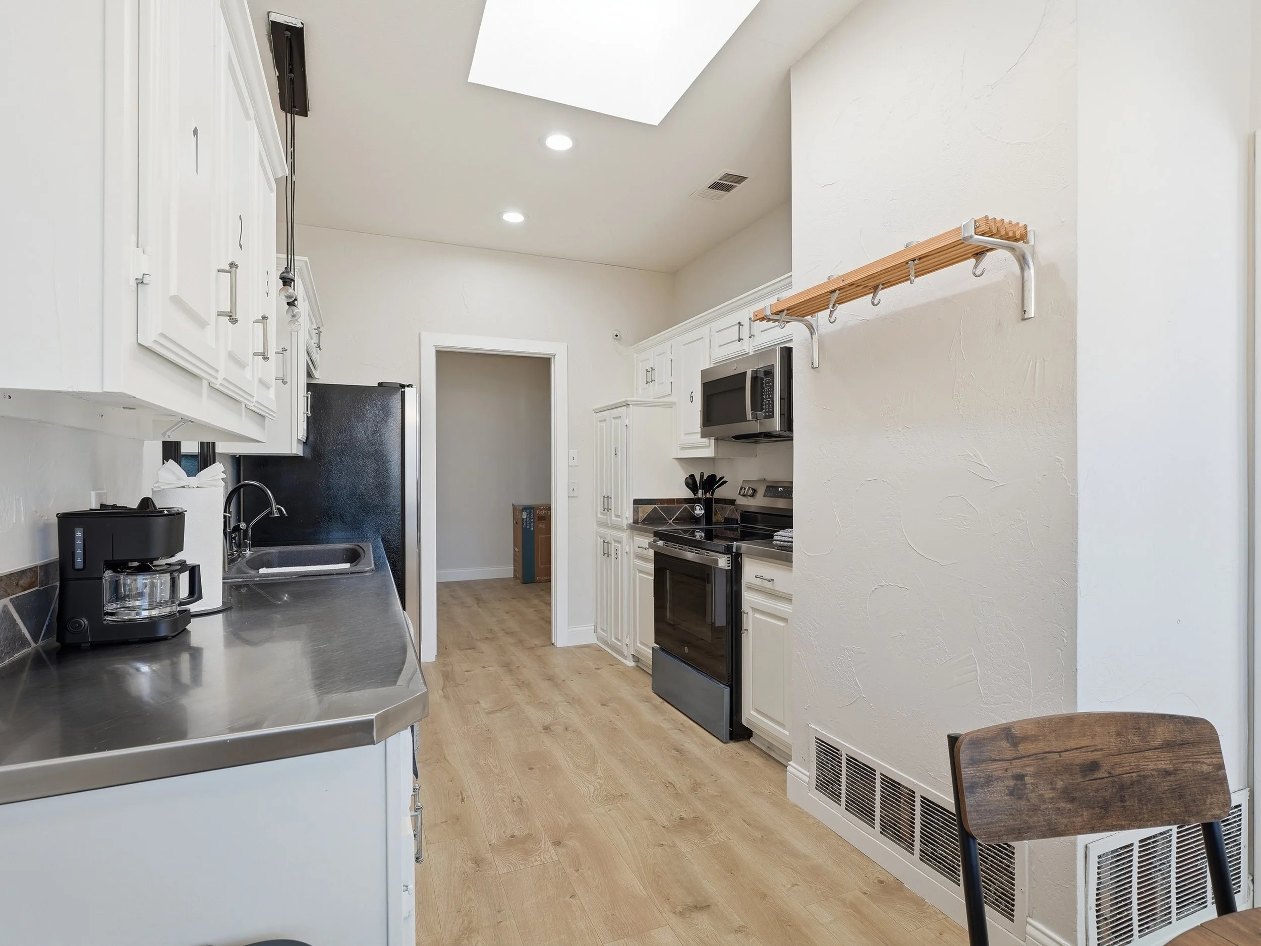Modern kitchen with white cabinets, stainless steel countertop, black appliances, oak flooring, and a wooden chair.