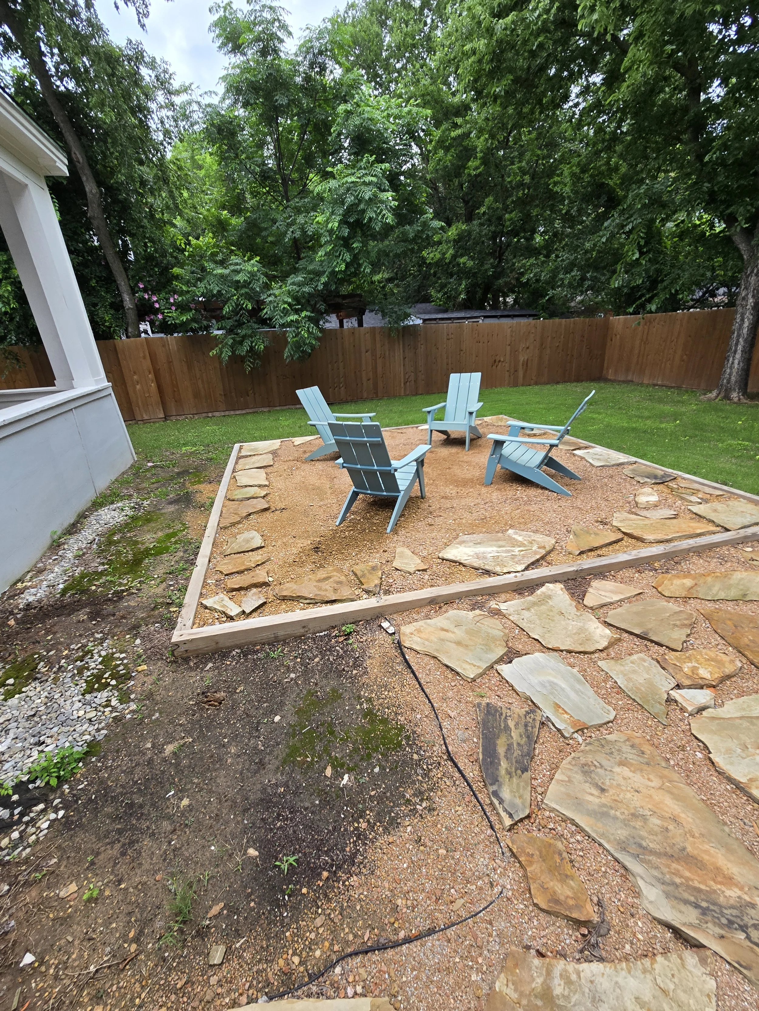 Backyard patio area with four blue Adirondack chairs, surrounded by a wooden fence, trees, and grassy lawn. Stone pathway leading into the patio.