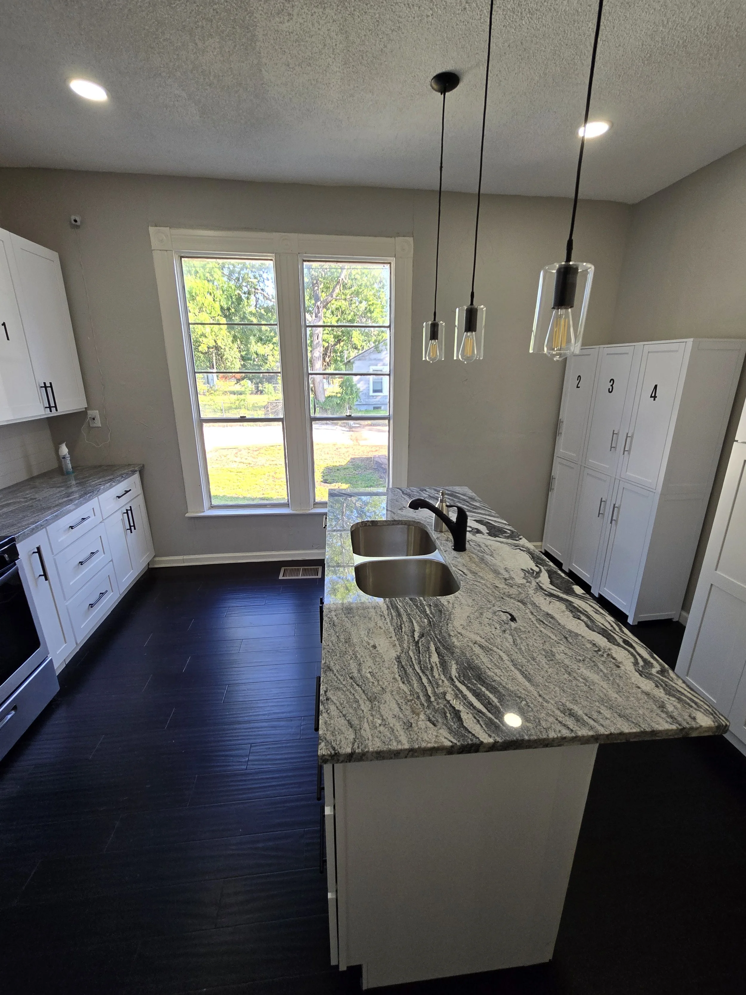 Modern kitchen with white cabinets, granite countertop, dark wood flooring, a large window, and pendant lights above the island.