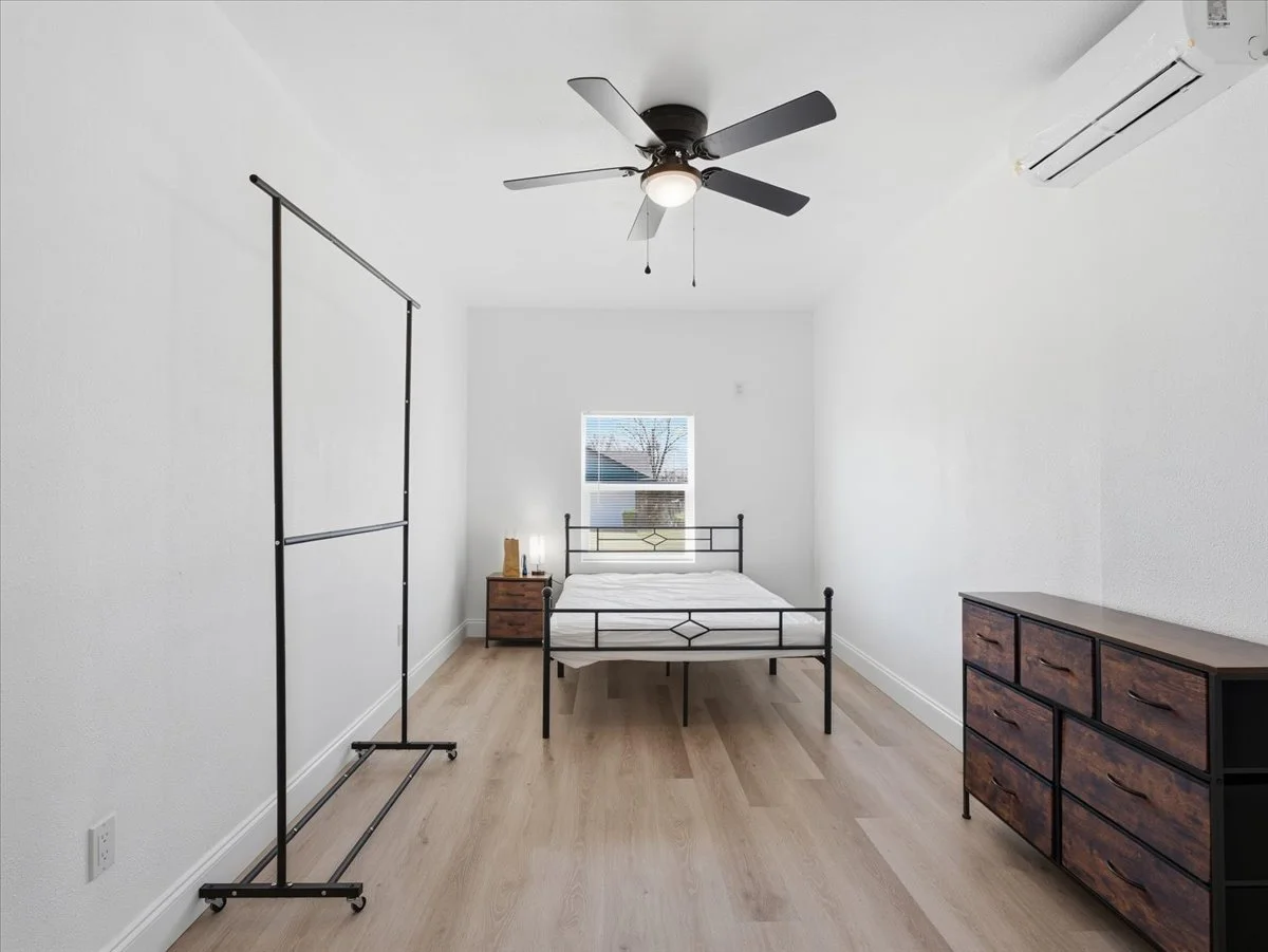 A minimalist bedroom with a bed, side table, dresser, clothing rack, ceiling fan, air conditioning unit, and window with blinds.