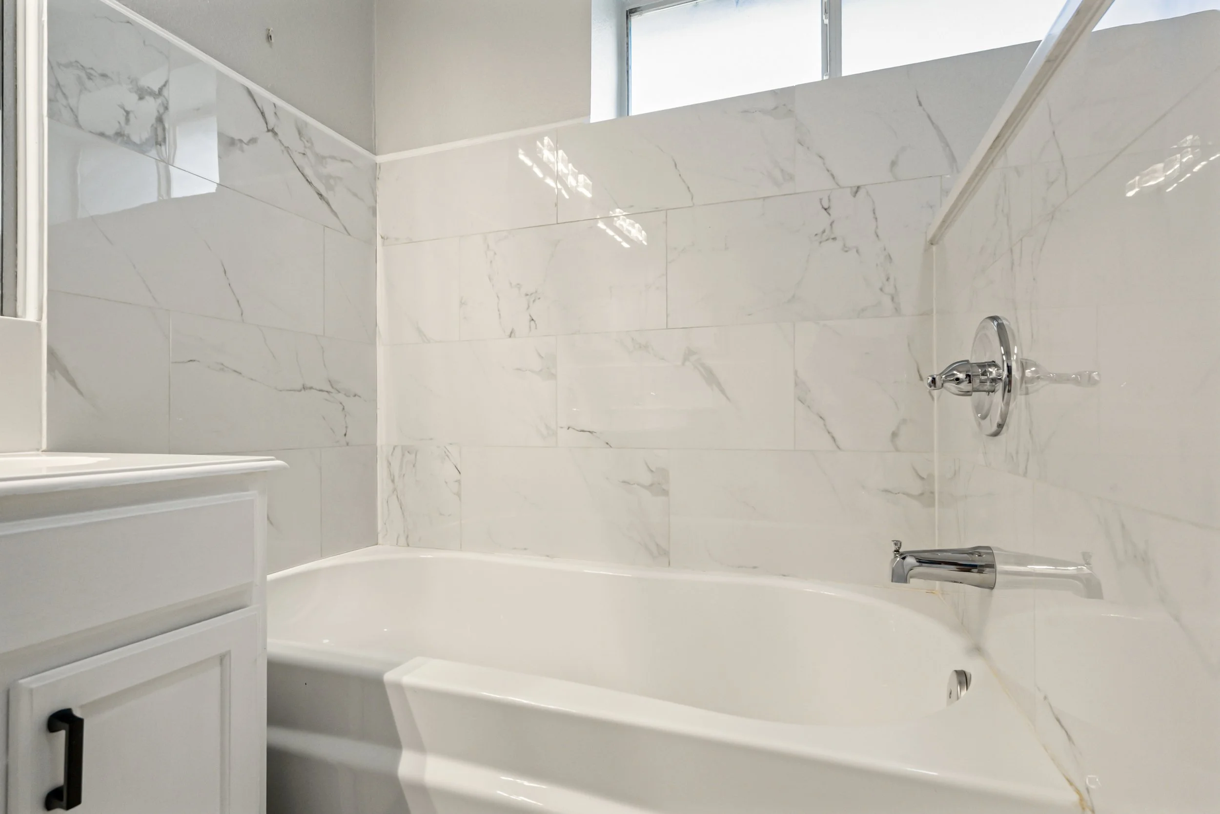 Bathroom with white marble tiles, a bathtub, silver fixtures, and a window above the shower area.