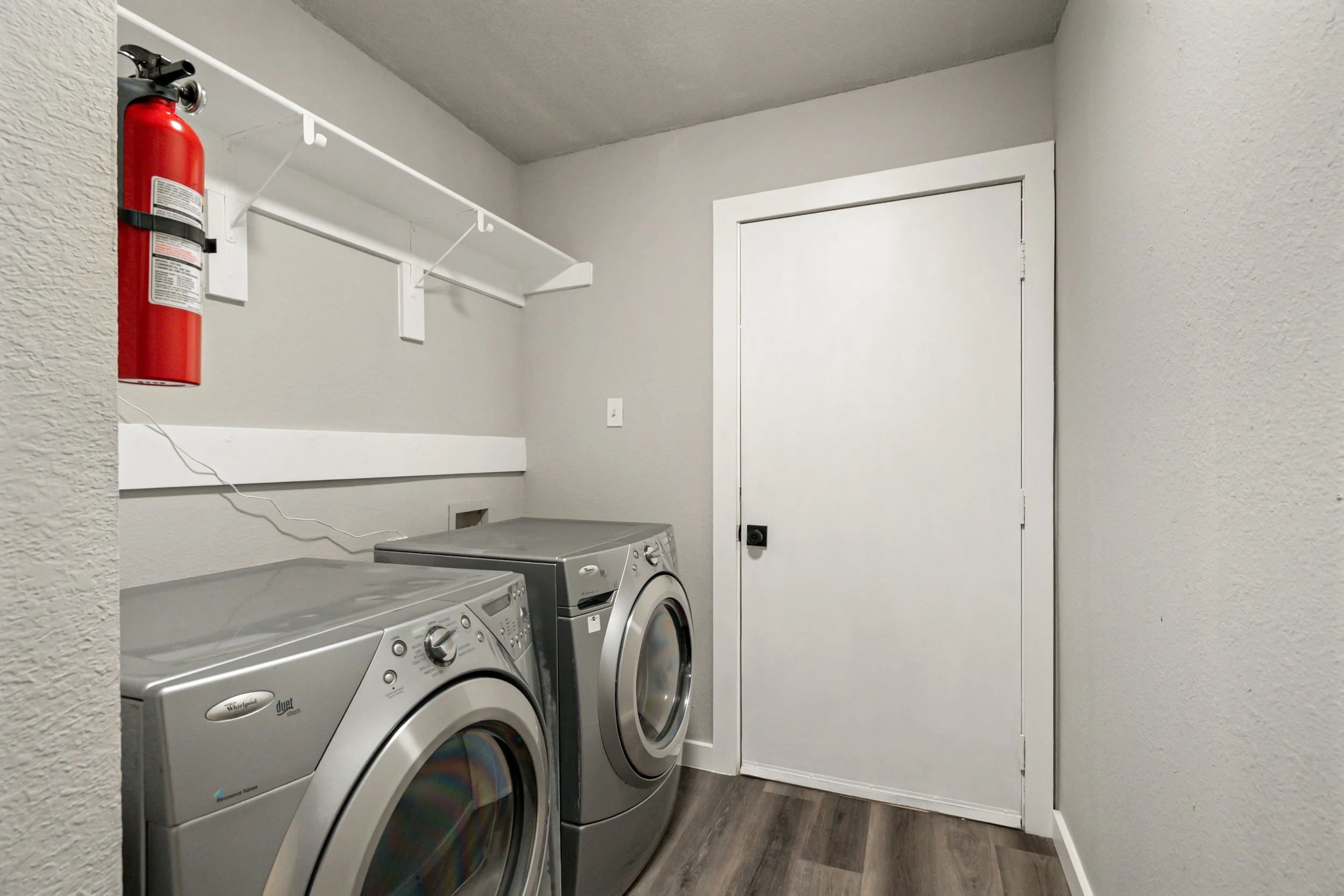 A laundry room with a white door, grey walls, a fire extinguisher mounted on the wall, and a washer and dryer side by side on a wood-like floor.