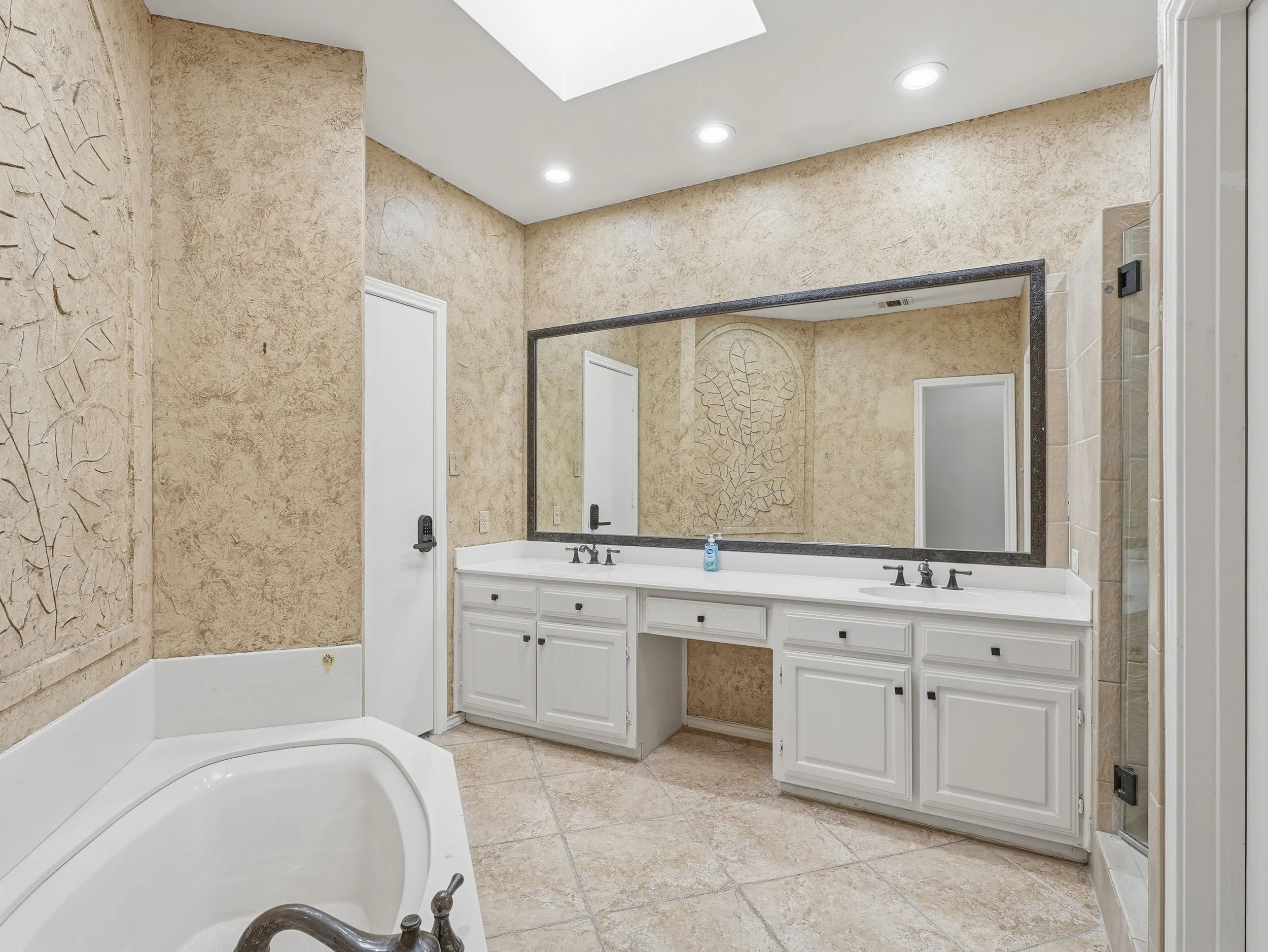 Bathroom with a large mirror above a white double sink vanity, a bathtub in the corner, and a shower stall on the side, with textured beige walls and tile flooring.