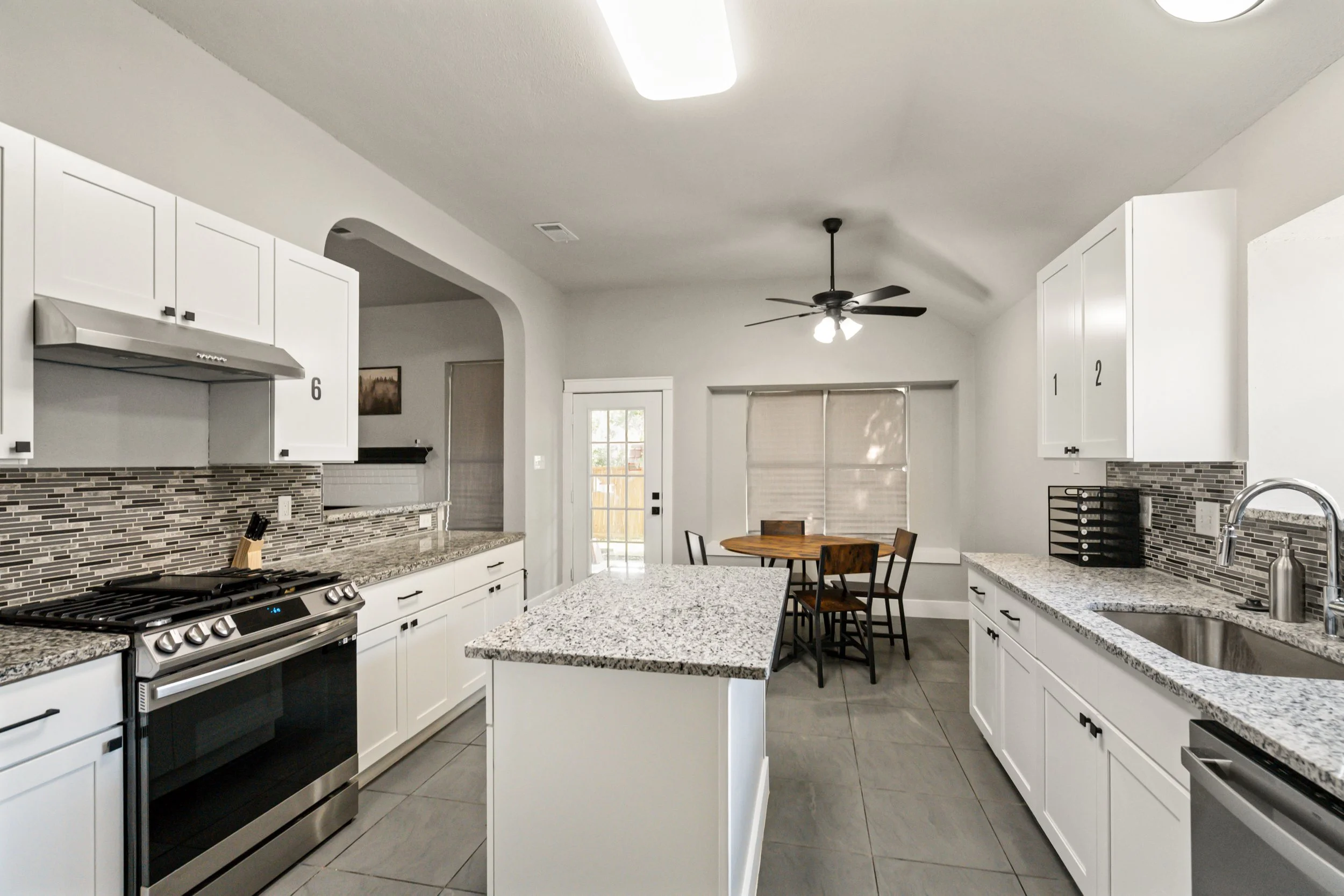 Modern kitchen with white cabinets, granite countertops, stainless steel appliances, tiled backsplash, island, and a dining area with a wooden table and chairs, ceiling fan, and window with blinds.