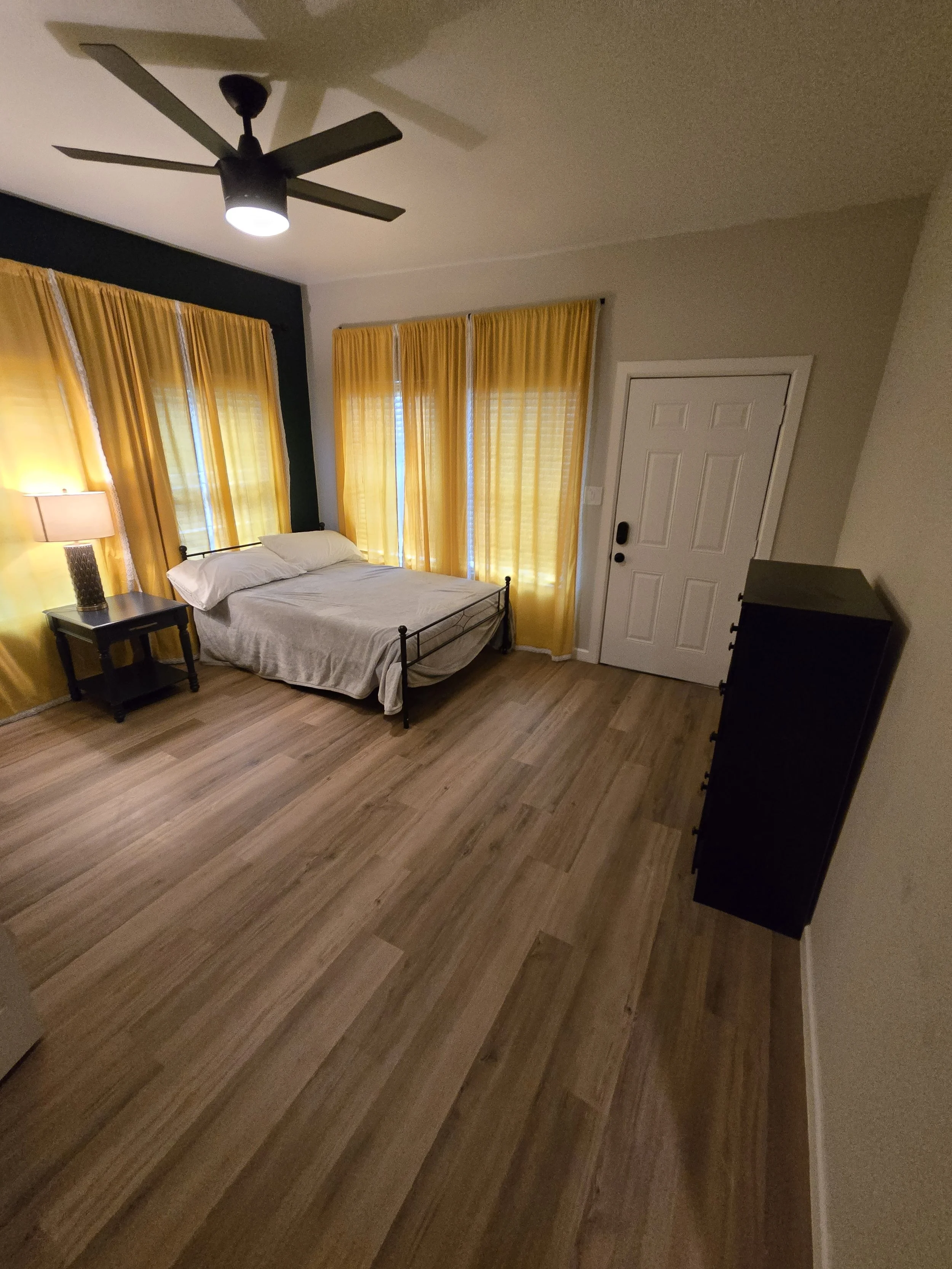 A bedroom with yellow curtains, a bed with white sheets, a black nightstand with a lamp, a black dresser, and a ceiling fan.