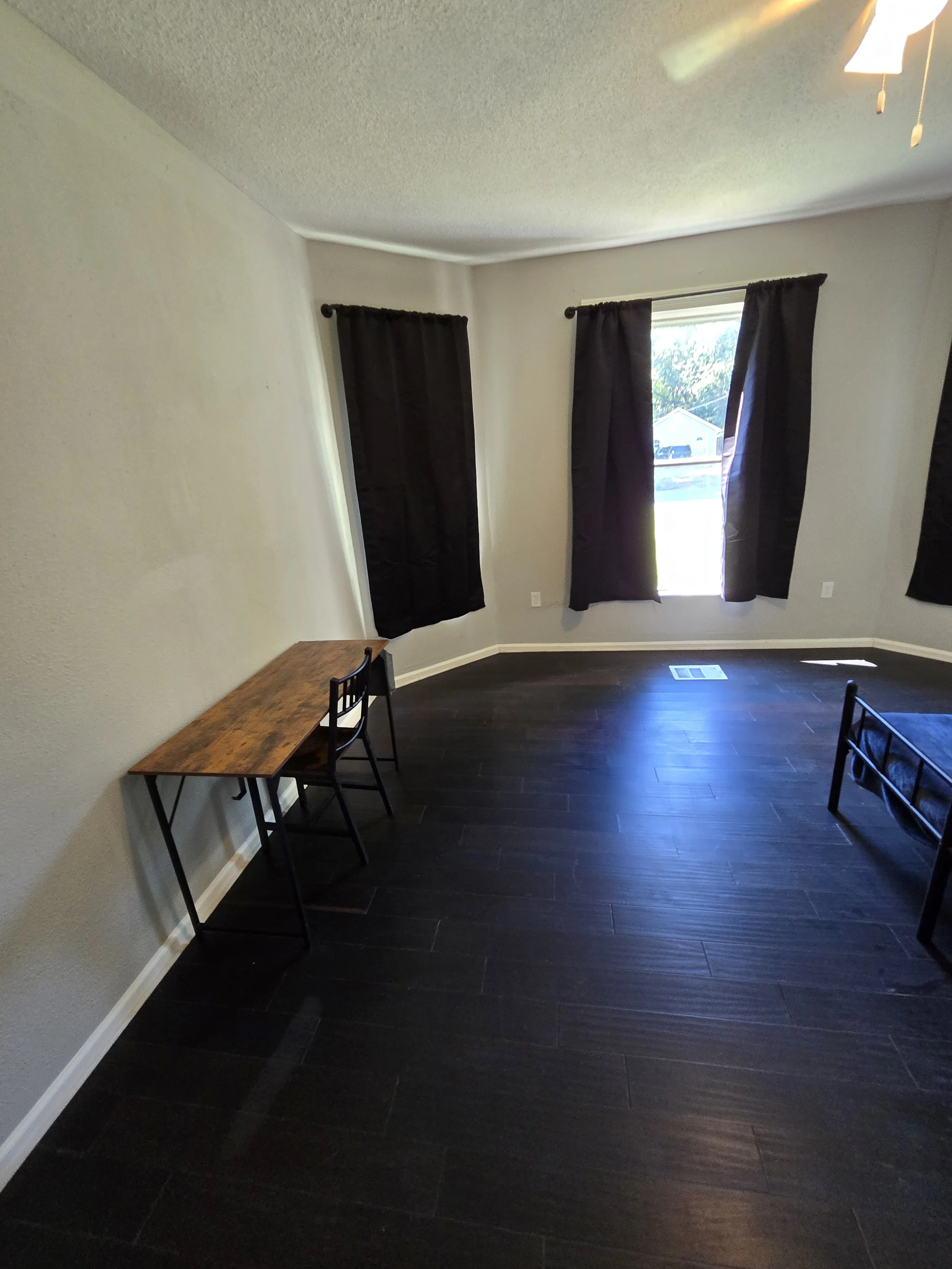 Empty bedroom with dark wood flooring, beige walls, and a ceiling fan. Two windows with black curtains, a small wooden desk with two chairs, and a partially visible bed frame.