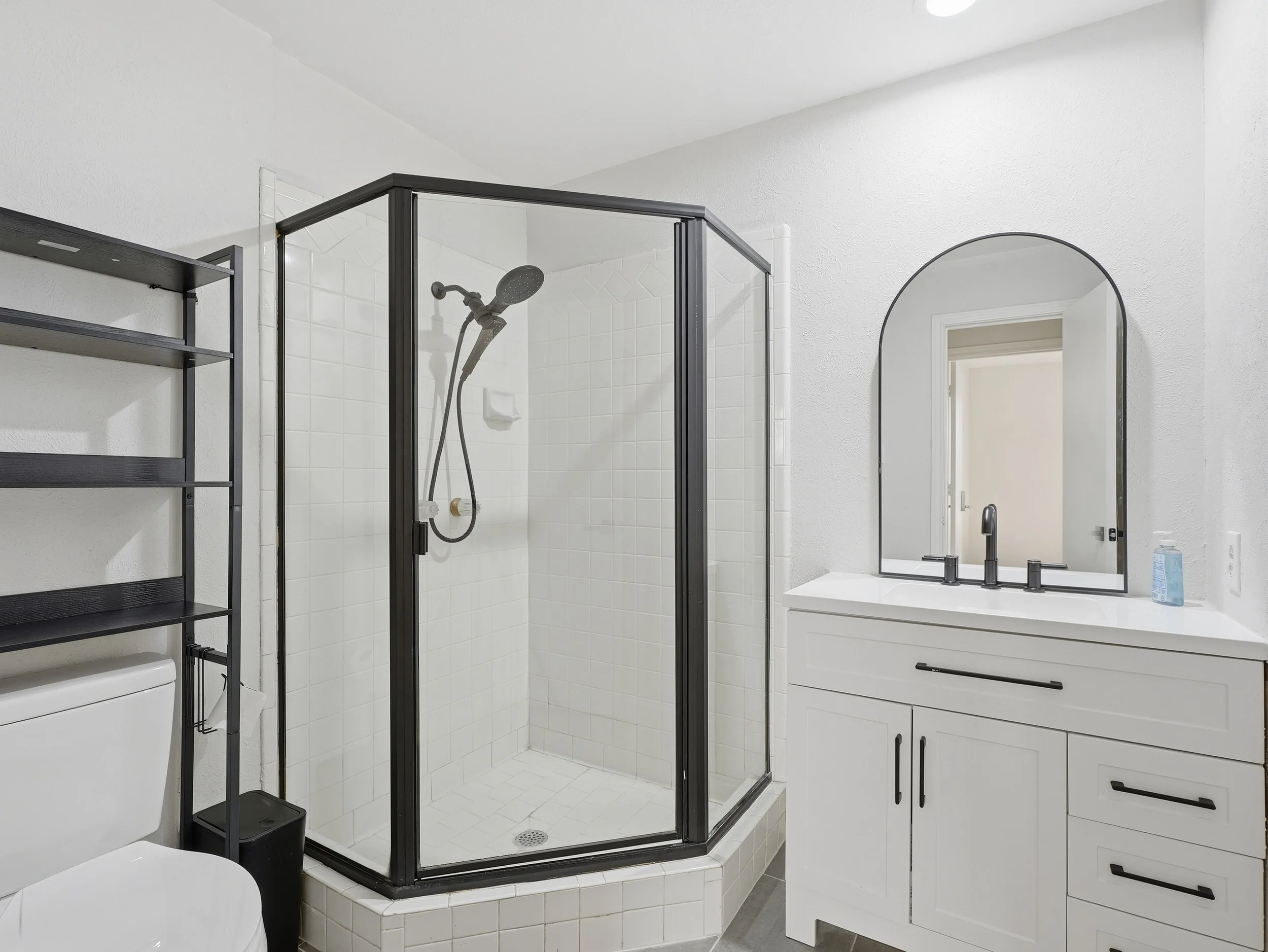 Bathroom with a glass shower enclosure, white tiled walls, a white vanity with a mirror, a hand soap bottle, and a black shelf above a toilet.