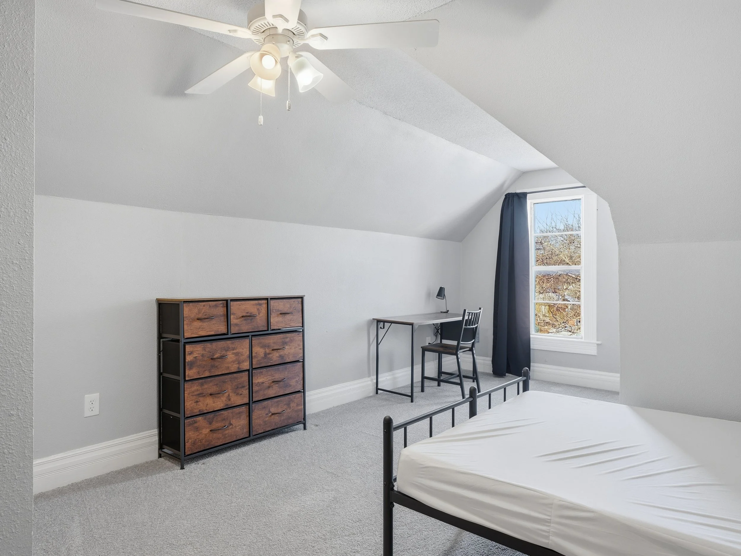 Empty bedroom with white walls and a sloped ceiling, featuring a metal bed frame with a mattress, a small wooden dresser, a desk with a lamp, a black chair, and a window with a dark curtain showing trees outside.